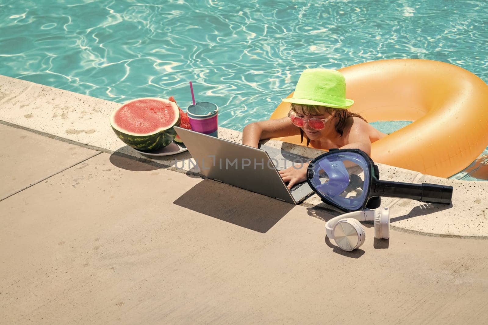 Child relaxing in the pool with laptop. Kid online working on laptop, swimming in a sunny turquoise water pool. Freelance work, distance online work, e-learning. by RedFoxStudio