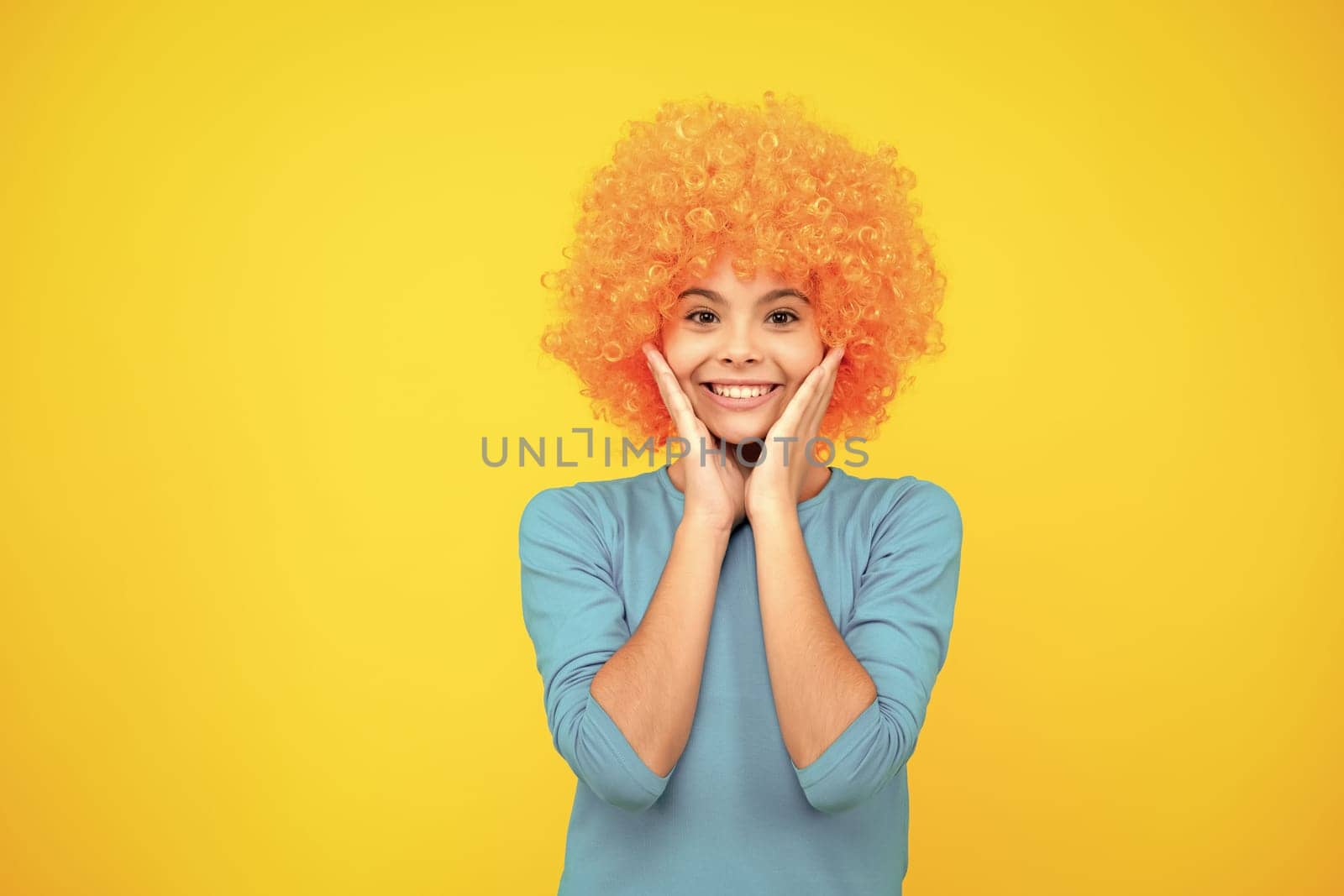 Birthday kids party. Funny kid in curly clown wig isolated on yellow background. Happy teenager portrait. Smiling girl