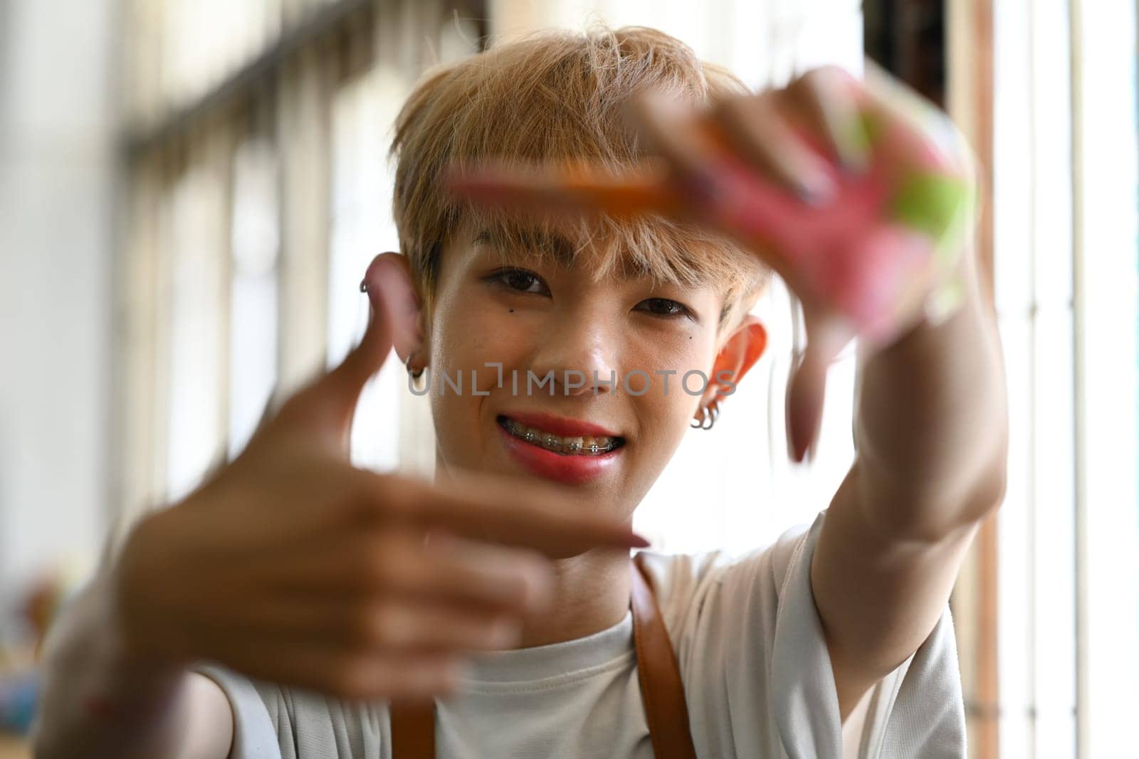 Happy young queer man framing his face with hands, looking at camera with a smile while standing in art studio by prathanchorruangsak