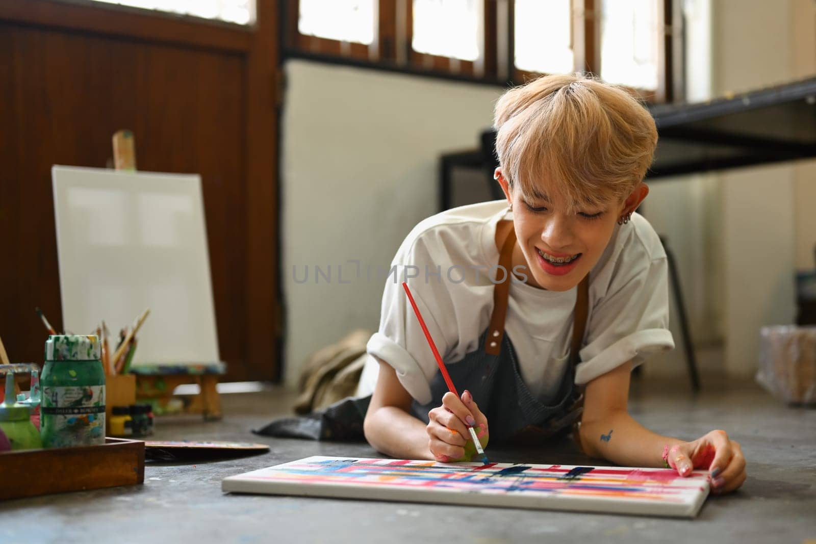 Carefree teenage male lying on floor in art studio and painting on canvas with watercolors. Creative hobby and art concept.