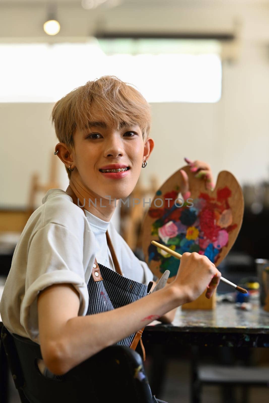 Cheerful young man art school student sitting in workshop holding palette and paintbrush, smiling at camera.