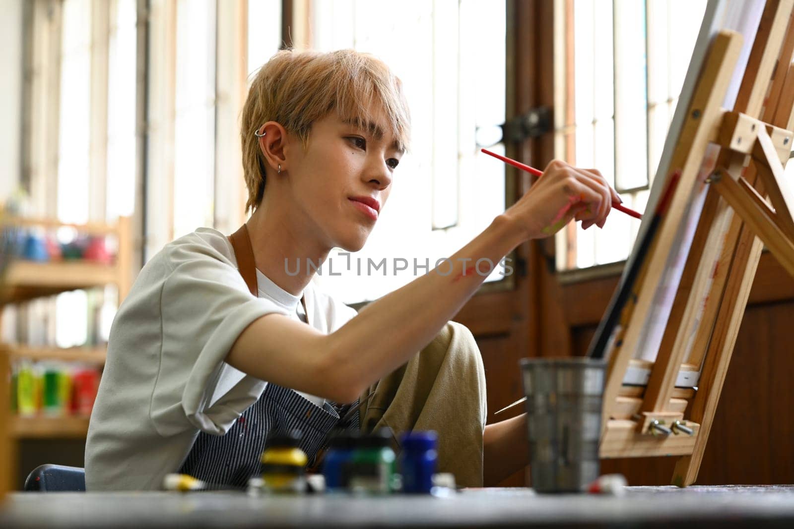 Concentrated asian student man in apron creating masterpiece on easel at art class. Art, education and creative activity concept by prathanchorruangsak