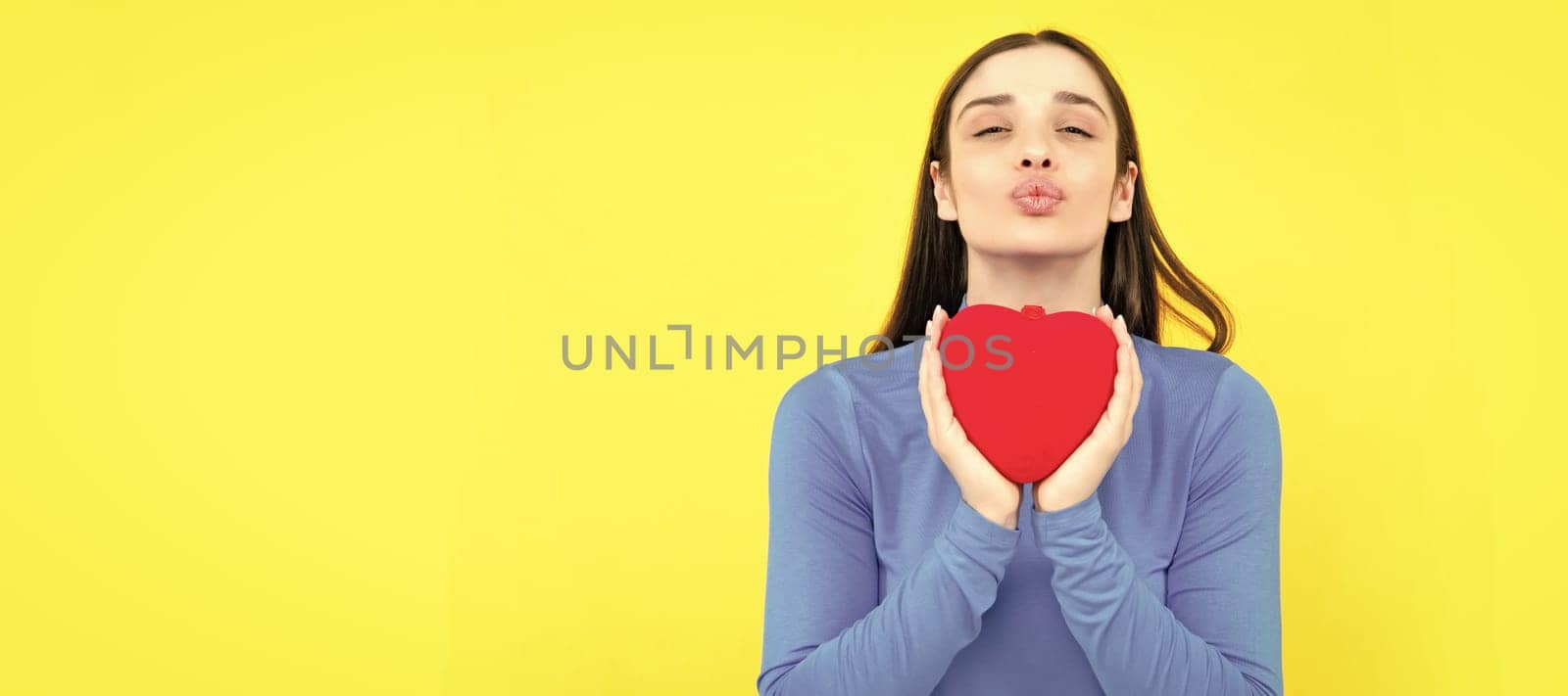 Beautiful girl holding valentines gift on yellow background. Portrait of young woman holding red paper heart. Woman isolated face portrait, banner with copy space. by RedFoxStudio