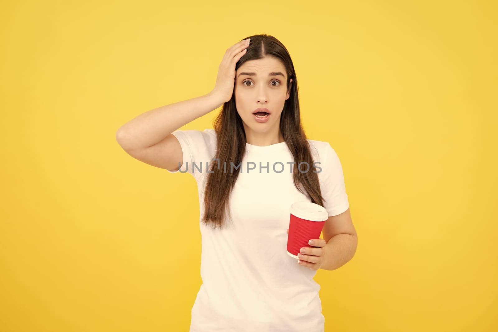 Close-up portrait of attractive dreamy girl holding in hands drinking latte isolated on yellow background. Young brunette woman drinking a cup of coffee