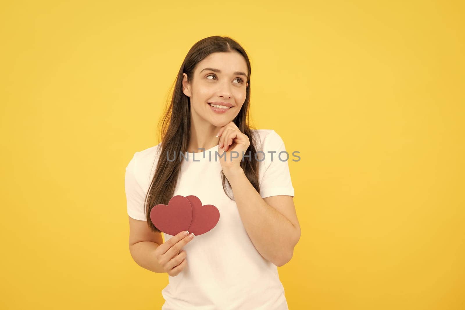 Romantic lovely girl. Portrait of attractive lovely cheerful girl holding in hands paper heart, isolated over yellow background. Love valentine concept