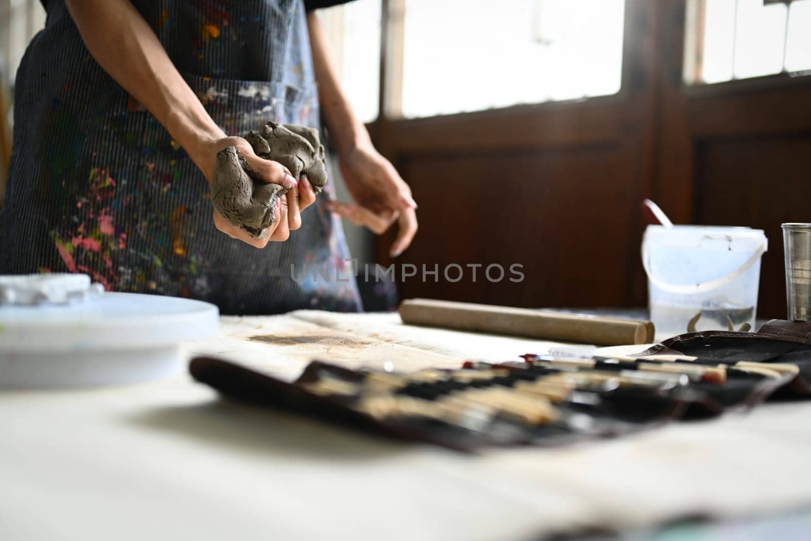 Pottery artist in apron kneading piece of raw clay with his hands on wooden table. Handicraft, creativity, hobby and activity concept by prathanchorruangsak