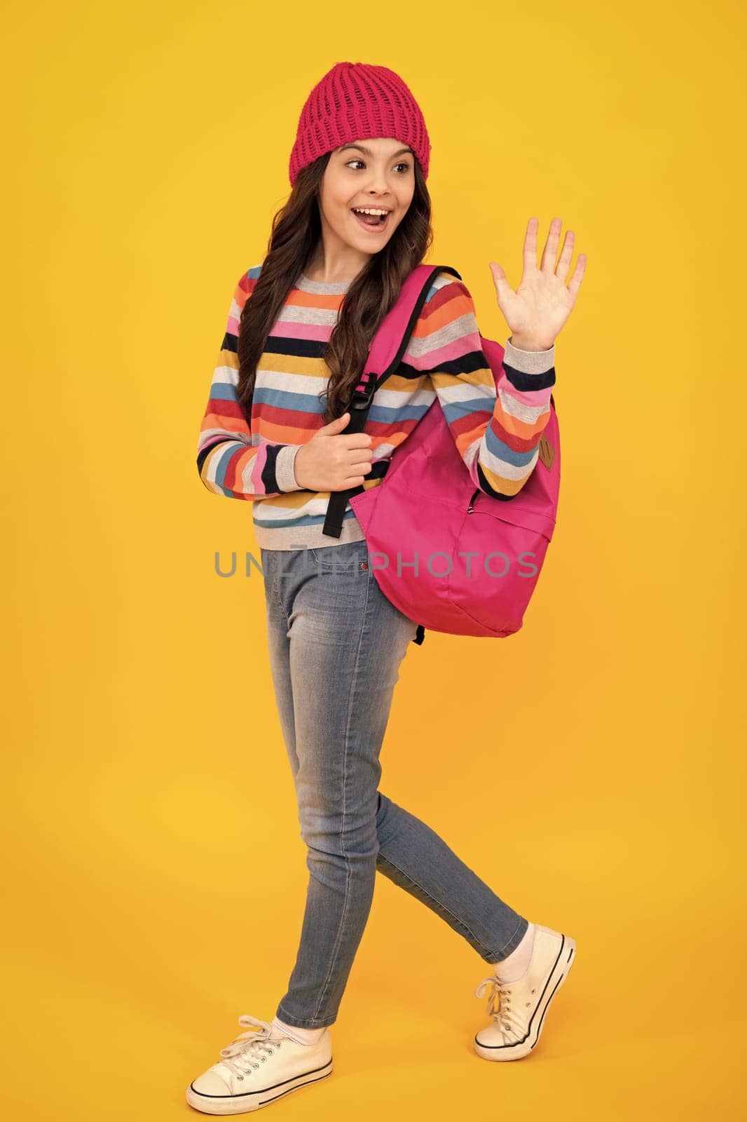 School children in winter hat and sweater with school bag on isolated yellow studio background. Children learning and education. Happy teenager, positive and smiling emotions of teen schoolgirl