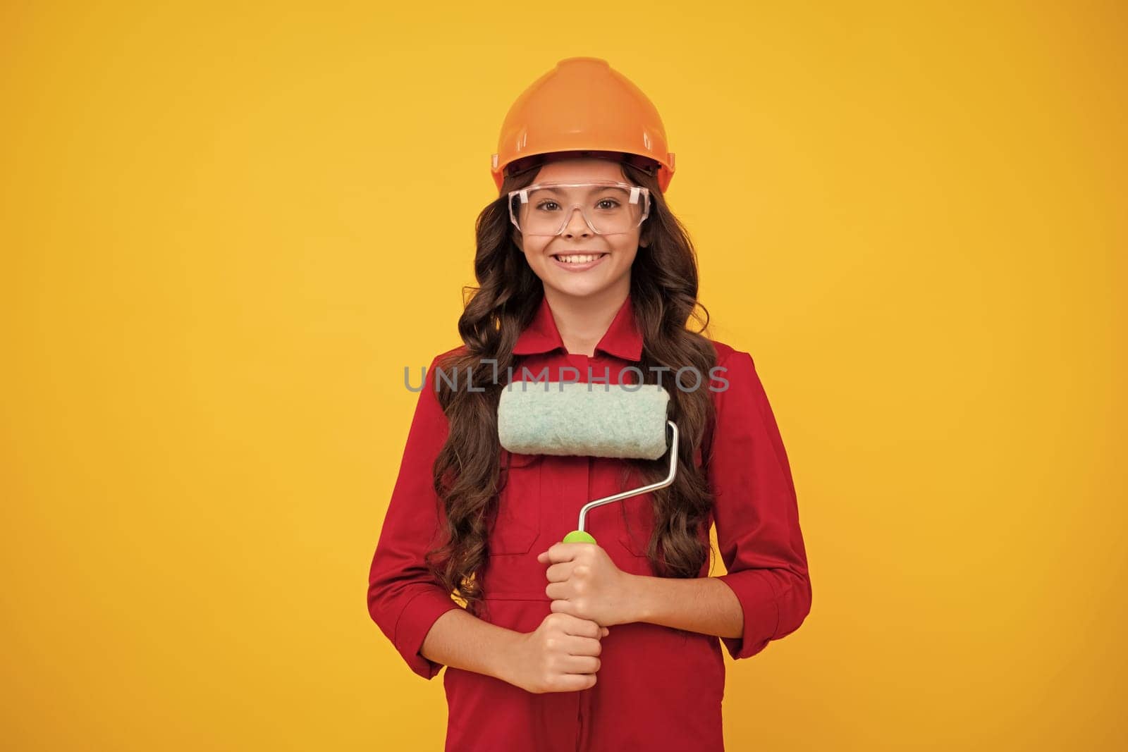 Building, construction and profession concept - smiling little girl in protective helmet and safety vest with paint roller over yellow background. Happy smiling teenager girl. by RedFoxStudio