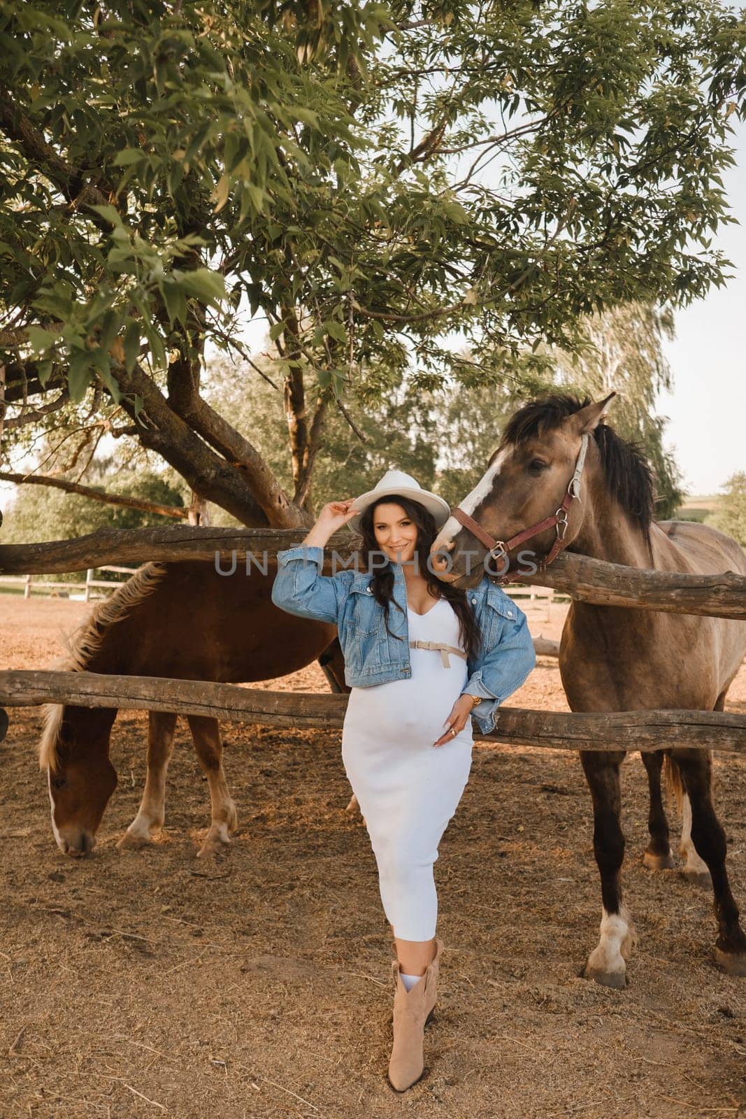 Stylish pregnant woman in a white hat in the countryside near a horse.