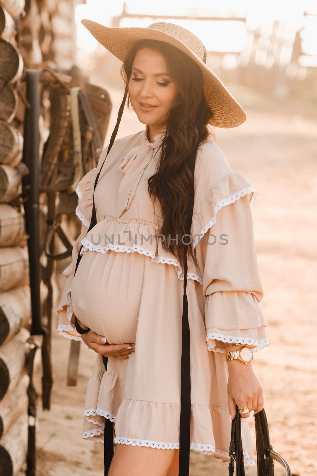 a pregnant woman in a dress and hat in the countryside holds a horse bridle in her hand.