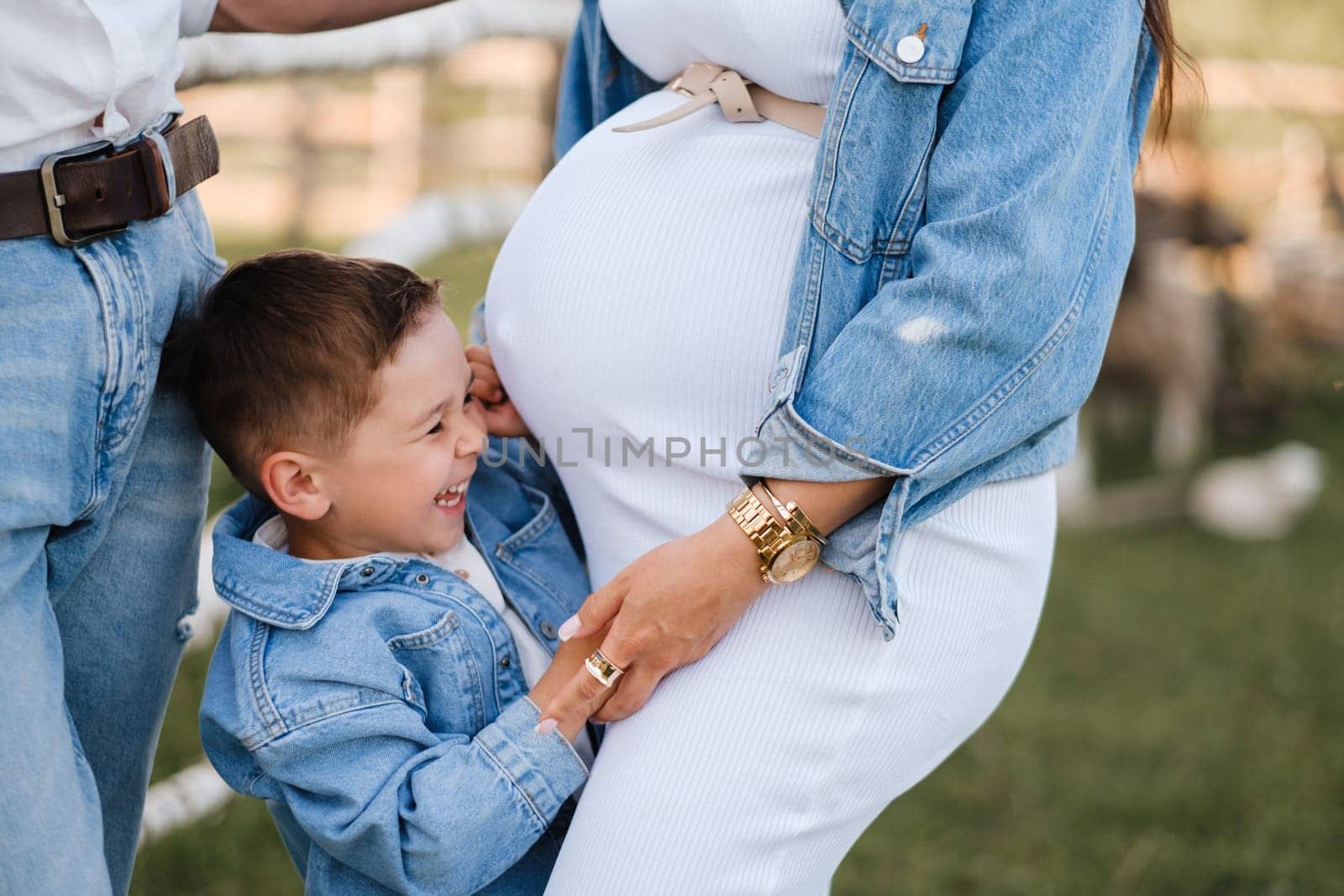 A little boy hugs the belly of a pregnant mother in a white dress in nature by Lobachad