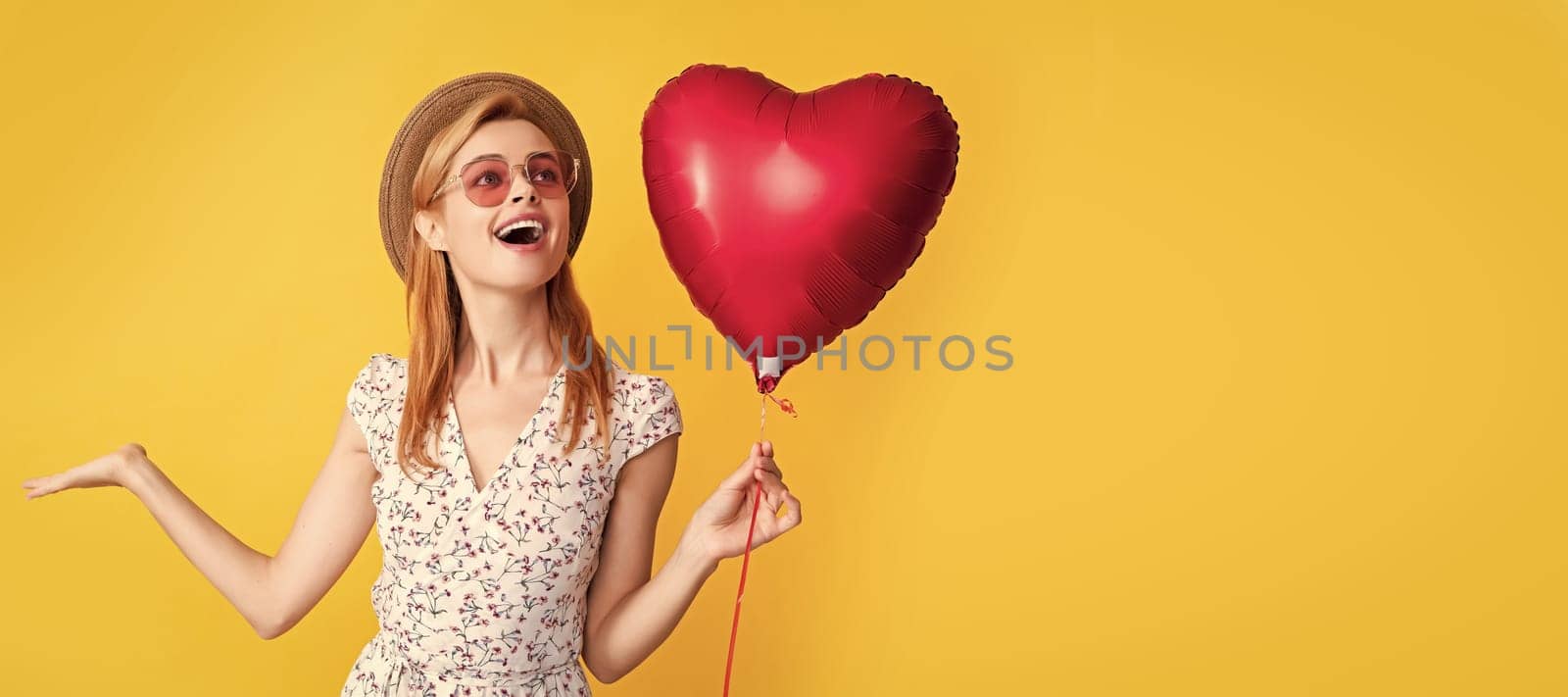 happy girl in straw hat and sunglasses hold love heart balloon on yellow background. Woman isolated face portrait, banner with copy space
