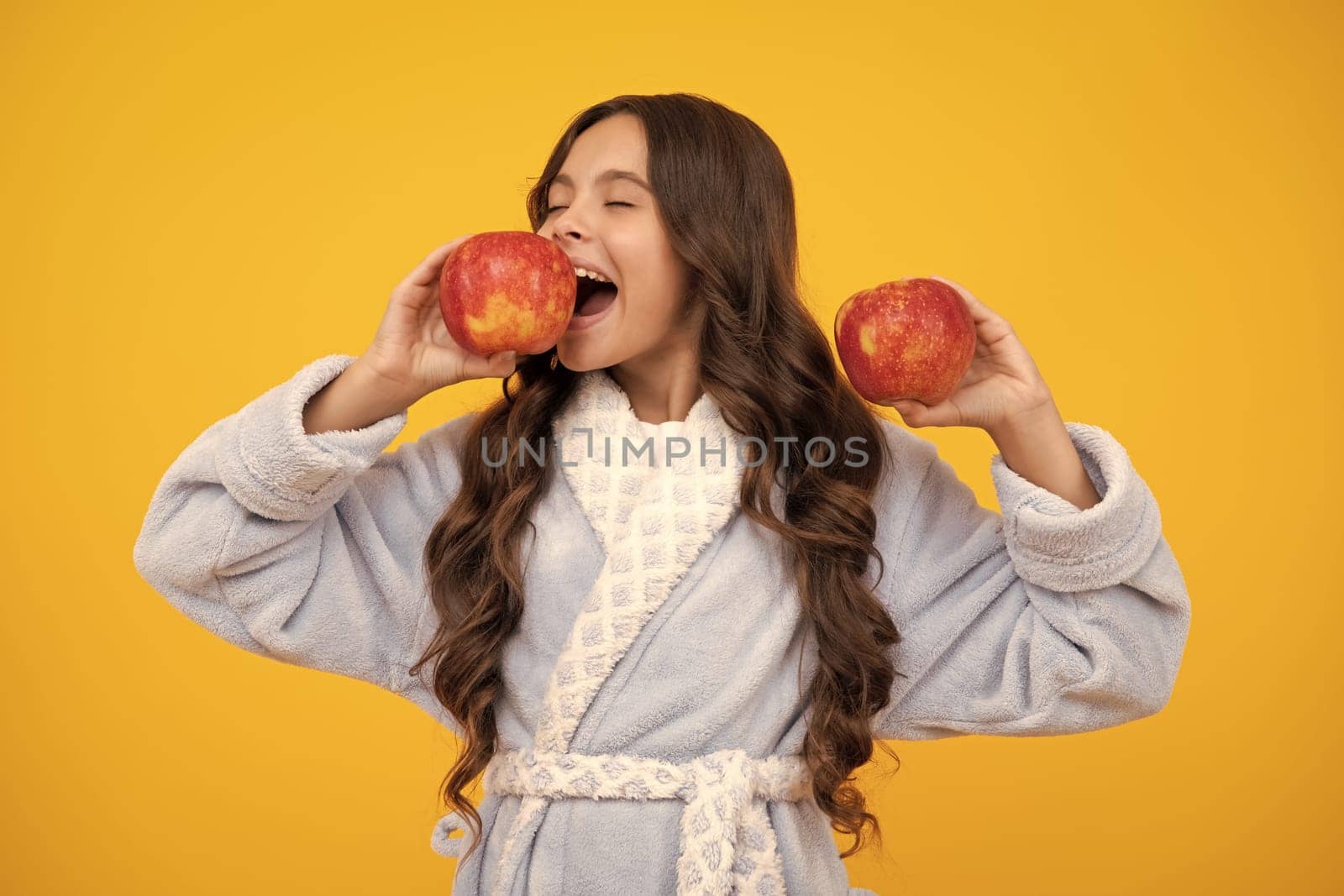 Child girl eating an apple over isolated yellow background. Tennager with fruit. Portrait of happy funny smiling teenage child girl. Surprised teenager girl