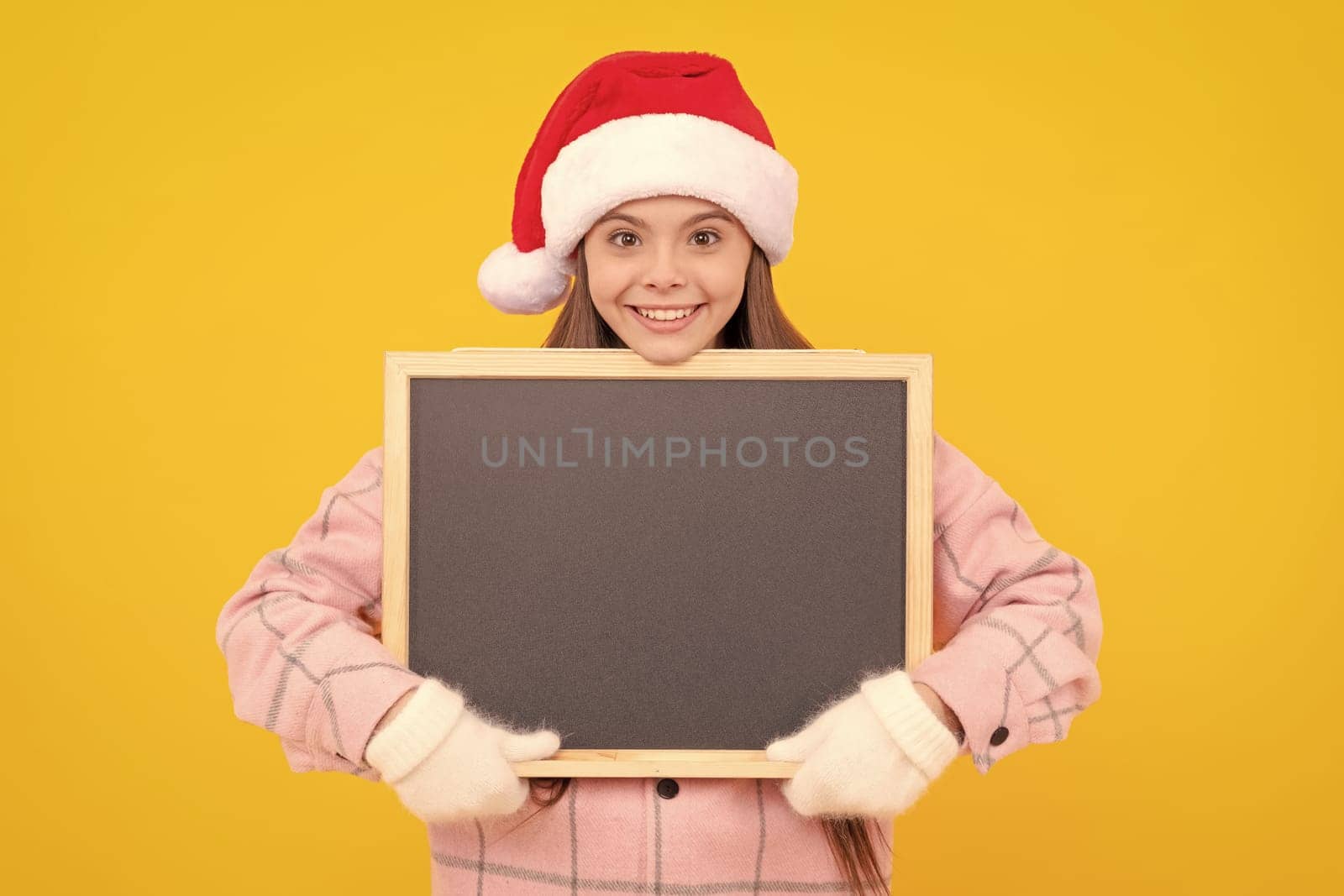 happy kid in santa claus hat hold blackboard for copy space. teen girl in mittens on yellow background. winter xmas holiday. child wearing warm clothes. back to school. happy new year. merry christmas