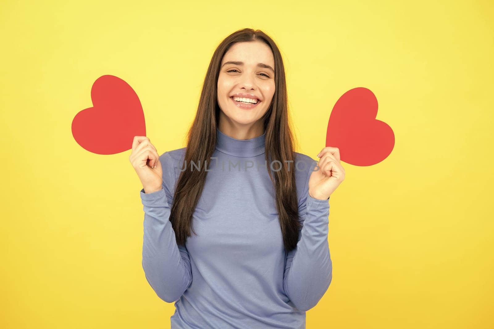 Beautiful girl holding valentines gift on yellow background. Portrait of young woman holding red paper heart