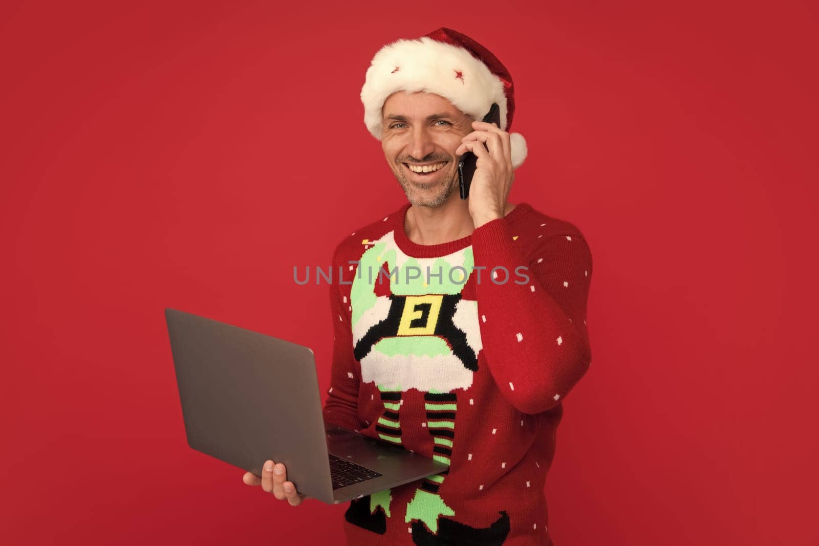 Santa with phone card and laptop. Man in Christmas sweater and hat on color background. Middle aged santa