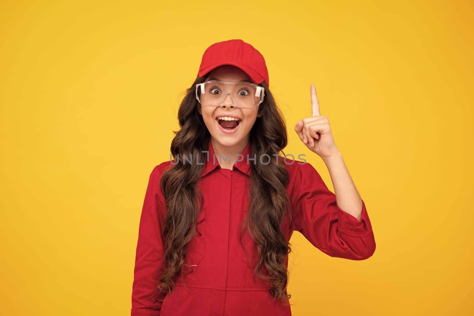 Worker teenager child wearing overalls red, cap and protect glasses. Studio shot portrait isolated on yellow background. Pointing and showing concept. Happy smiling emotions of teenager girl