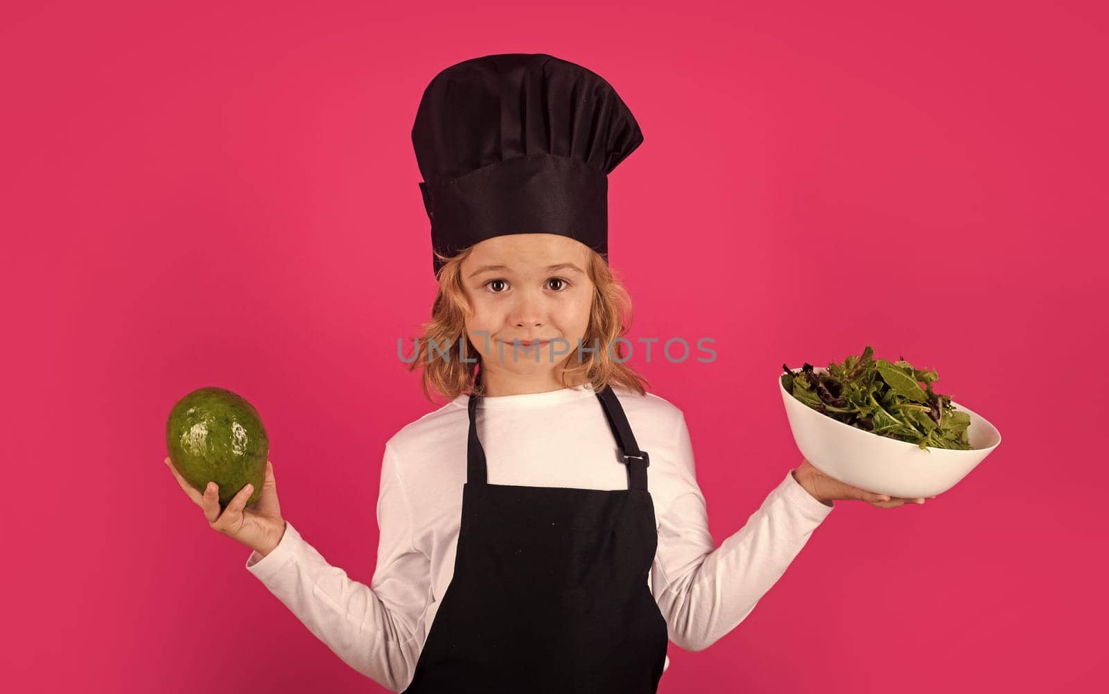 Funny child cook with avocado and vegetable. Child wearing cooker uniform and chef hat preparing food on kitchen, studio portrait. Cooking, culinary and kids food concept. by RedFoxStudio