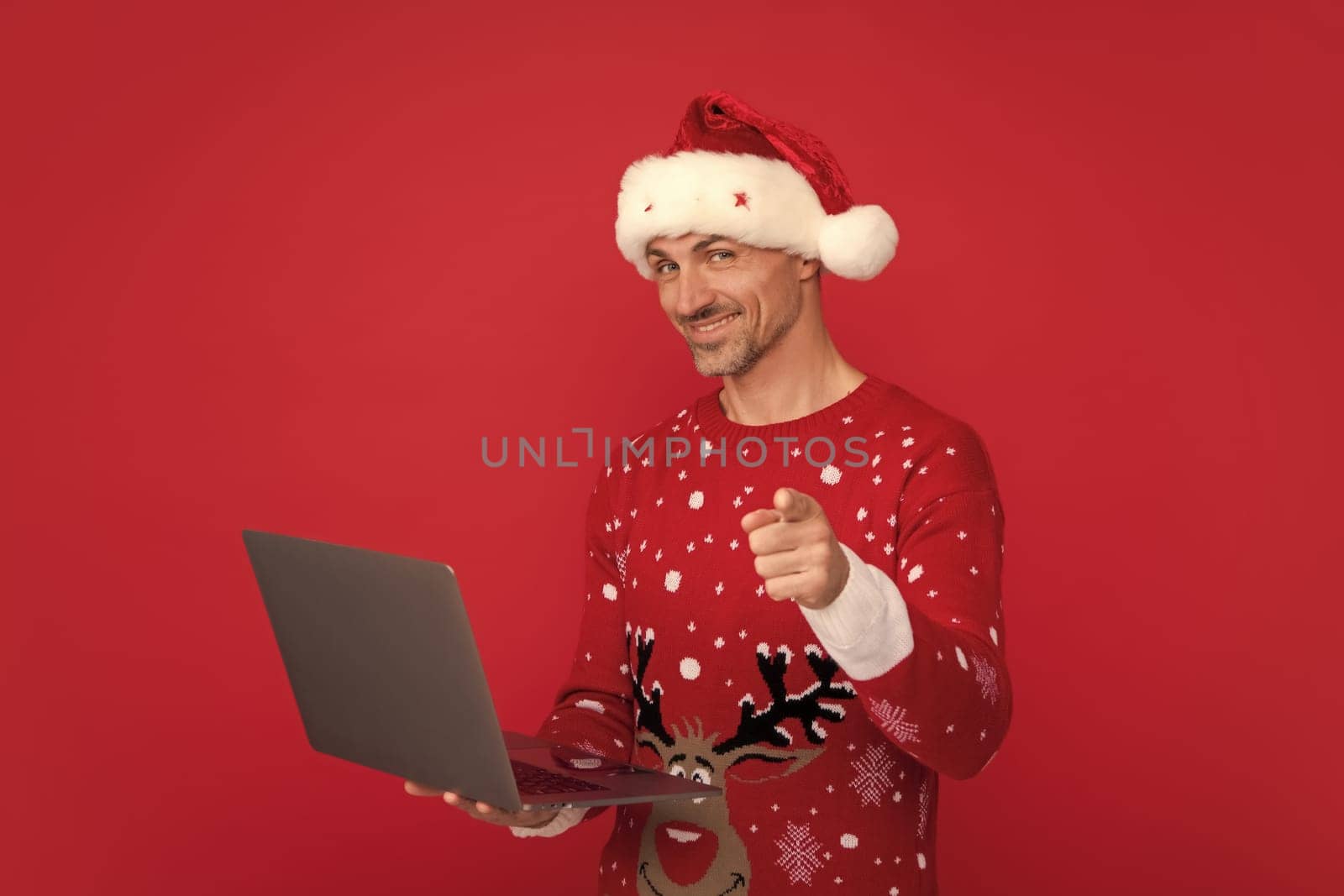 Santa with laptop, online shopping, winter business. Portrait of middle aged man in sweater isolated over red background. Concept of holidays, happiness, emotions and Christmas celebration