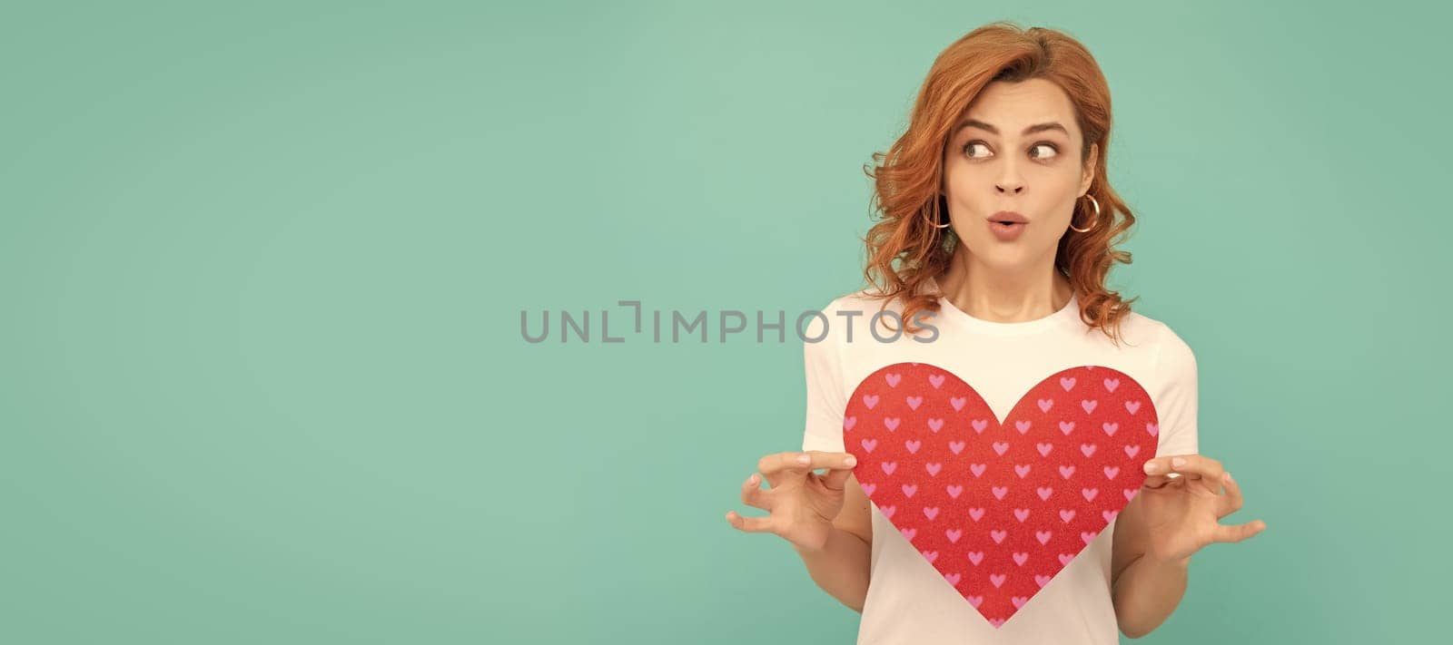 surprised redhead girl hold red heart on blue background. Woman isolated face portrait, banner with copy space