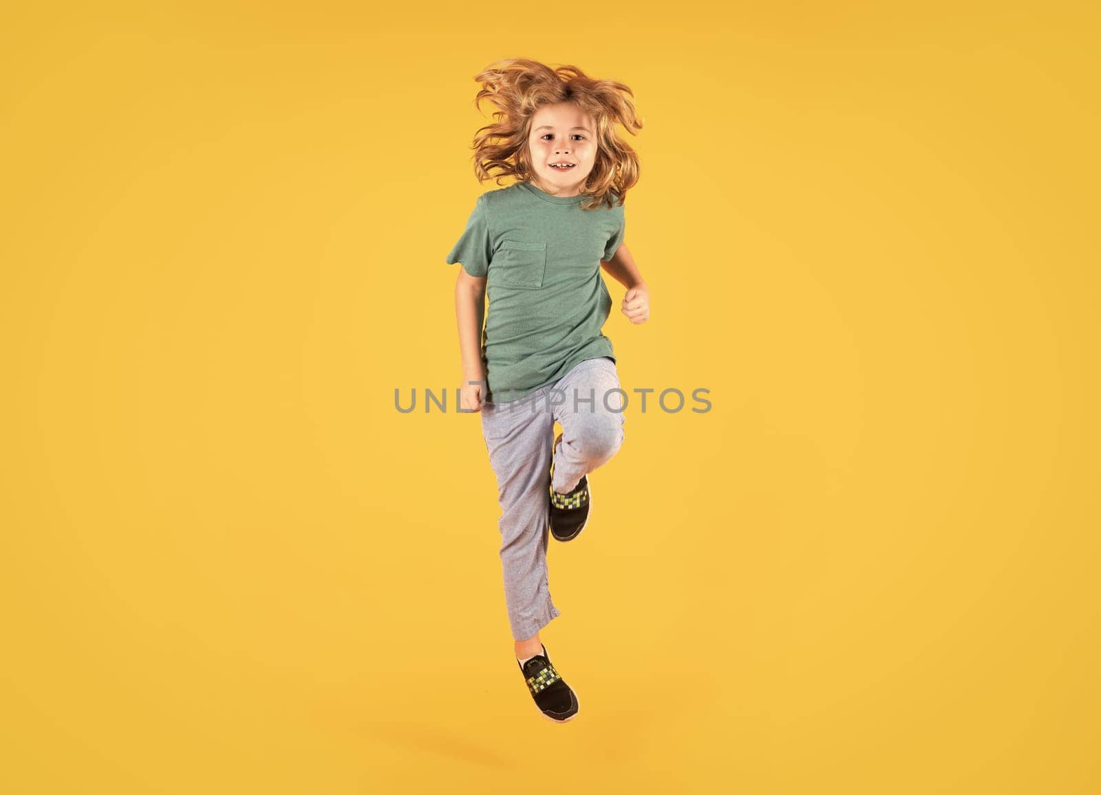 Energetic kid boy jumping and raising hands up on isolated studio background. Full length body size photo of jumping high child boy, hurrying up running fast on yellow background