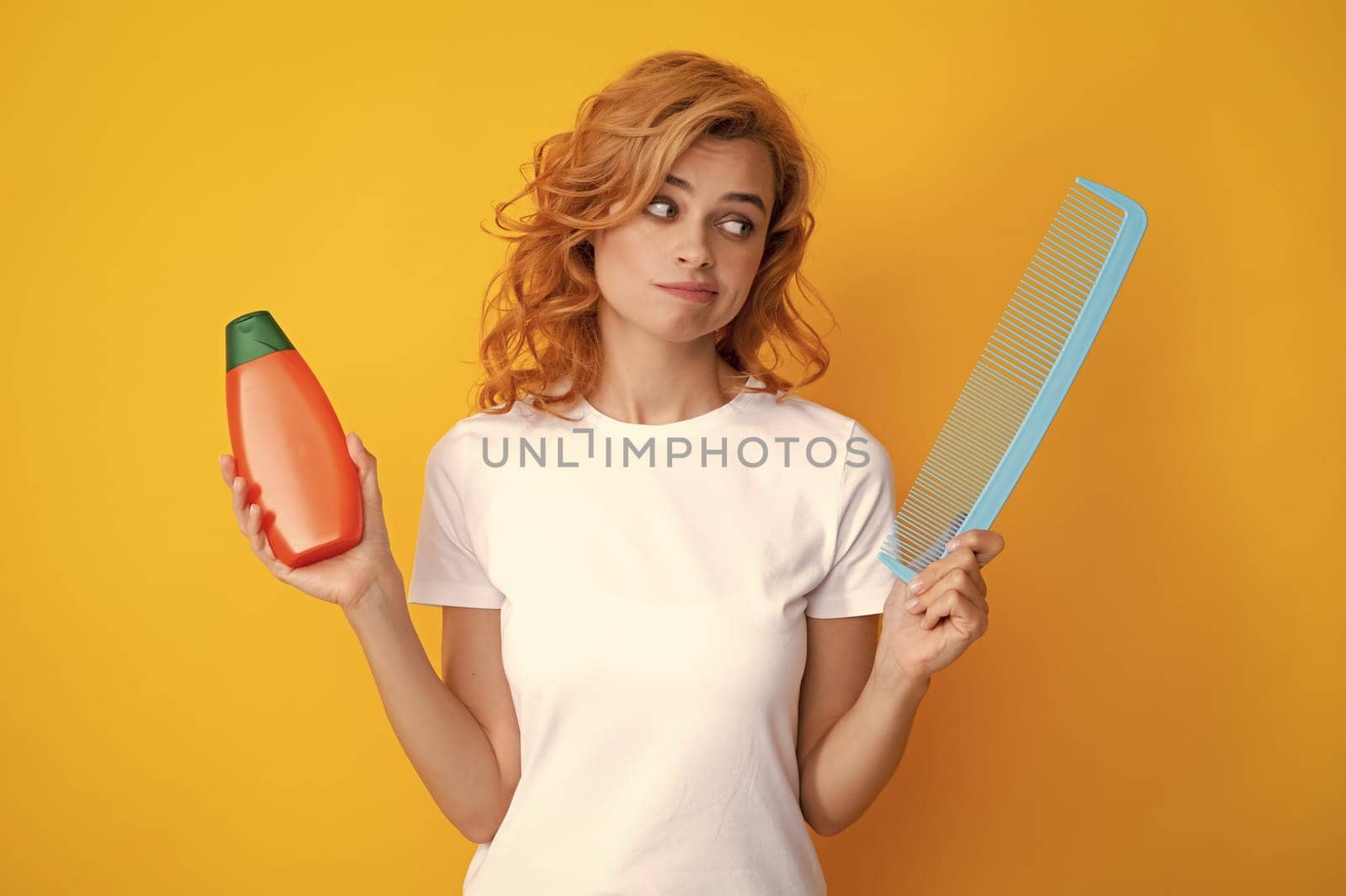 Angry woman hold bottle shampoo and conditioner. Redhead woman with a comb, isolated on yellow background