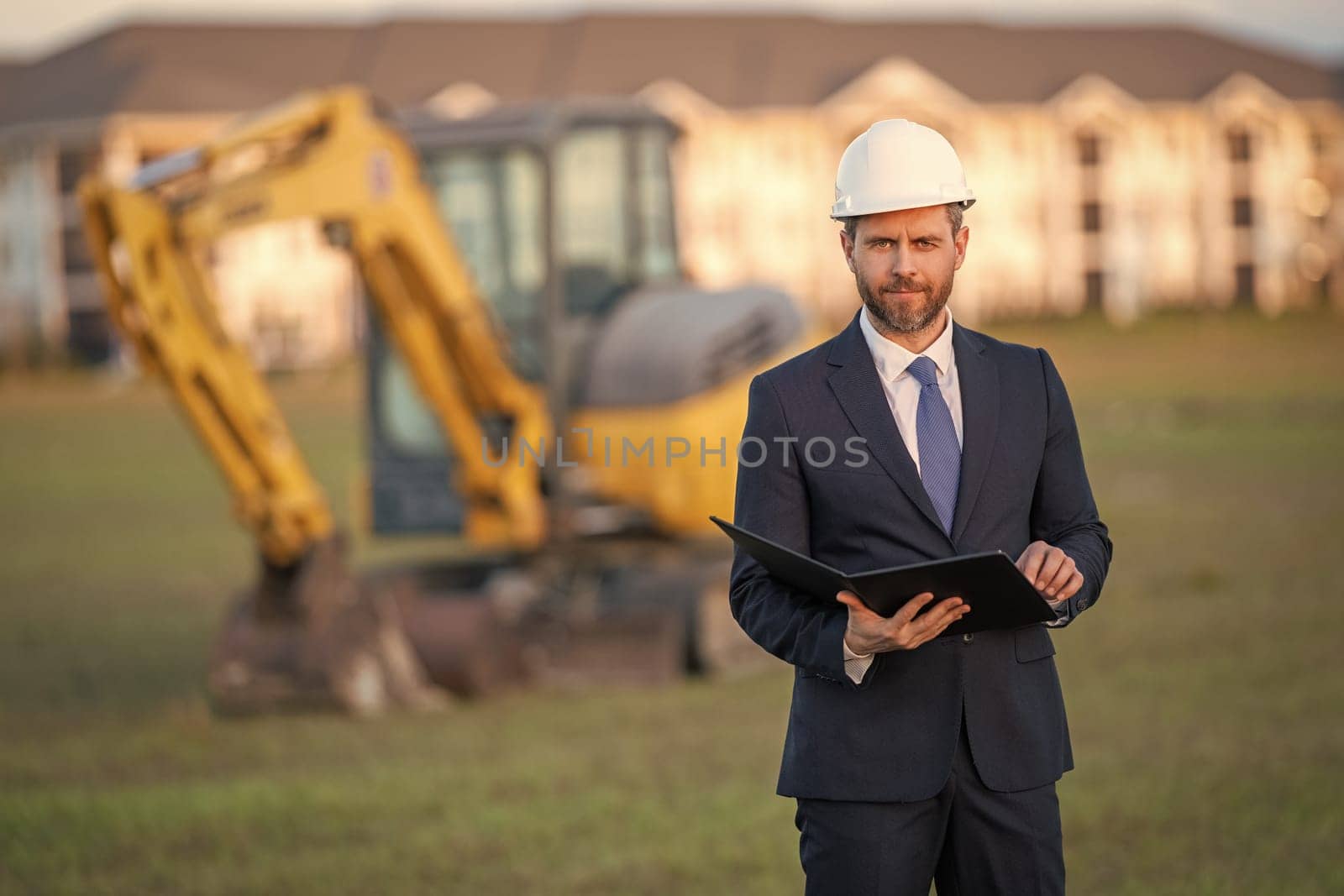 Architect at a construction site. Architect man in helmet and suit at modern home building construction. Architect with a safety vest and suit. Confident architect standing at house background