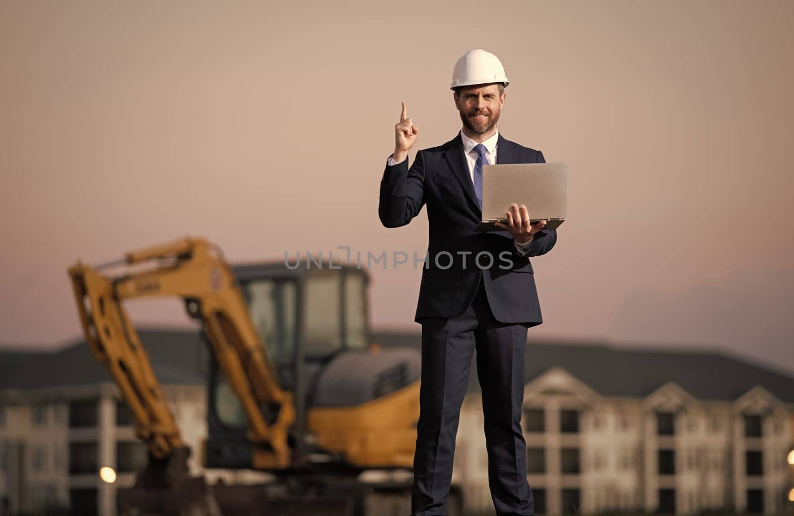 Buider man in suit and hardhat. Construction investor. Business man investor in front of construction site. Successful investor. Handsome man in suit and hardhat at building construction