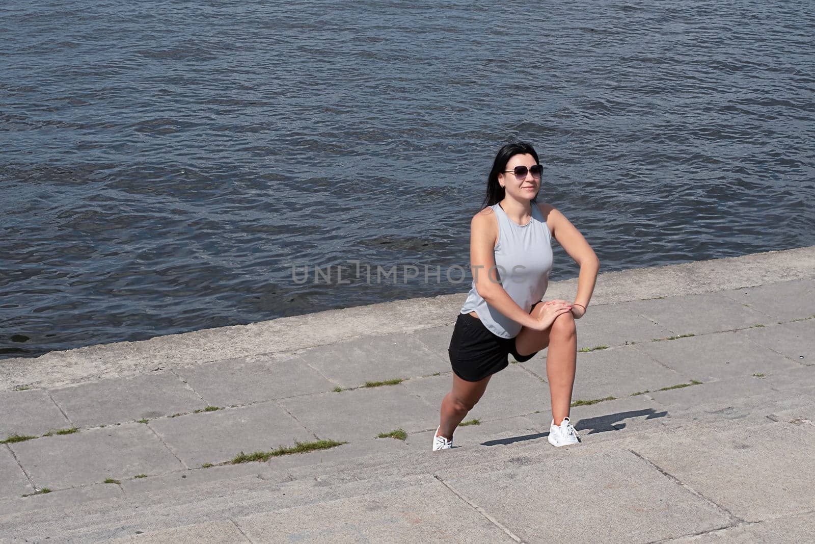 The girl is slender, tall and athletic in short shorts and sunglasses, in the summer she is engaged in fitness on the street by the river in the open air. Sports concept.