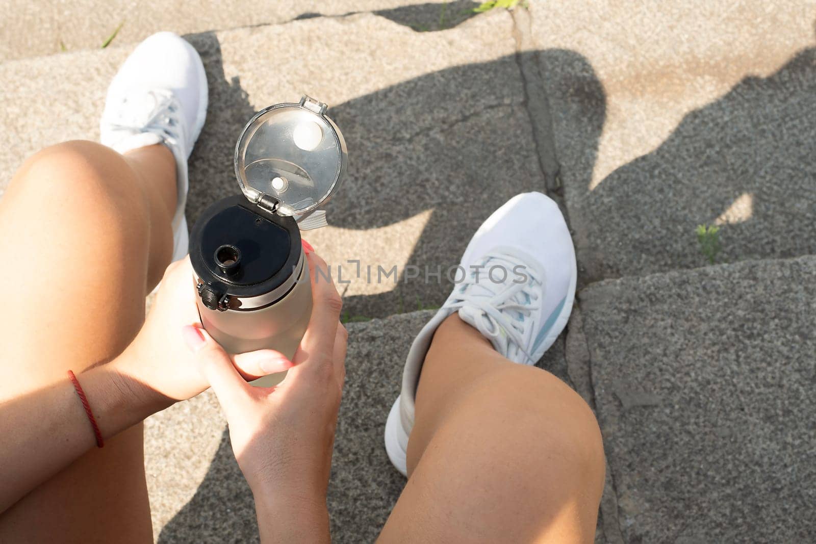 Sports concept. The girl sits on the steps in white sneakers and holds a bottle of water in her hand. Resting and quenches thirst after a workout on the street. No face. by ketlit