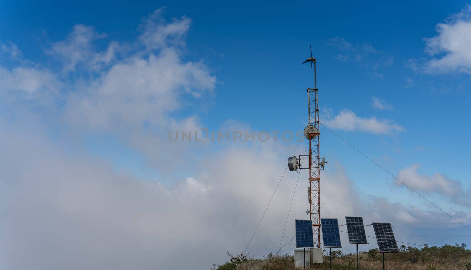 The communication antenna on top of the mountain is powered by solar panels and a small wind turbine, with a cloudy blue sky in the background.
