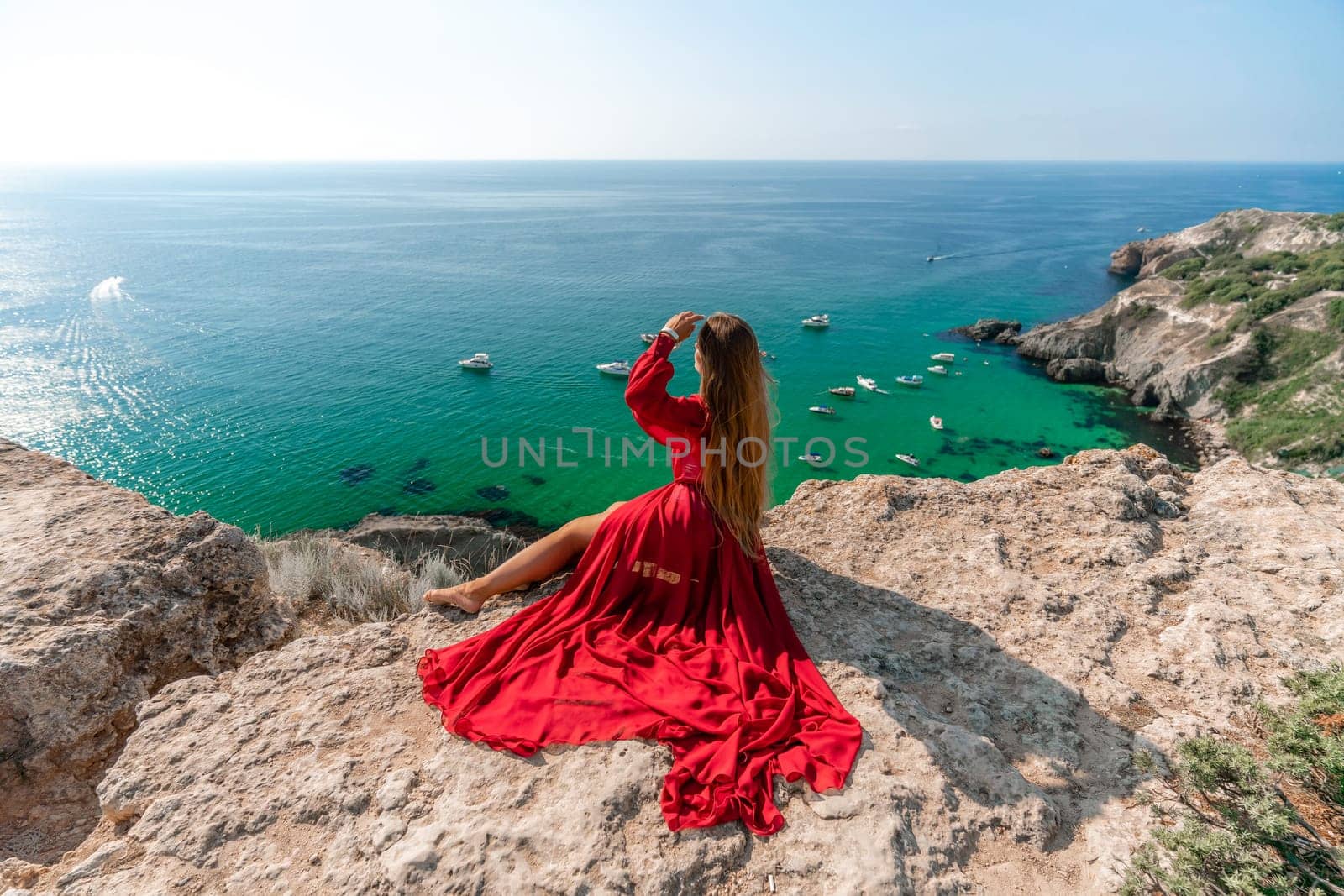 Woman red dress sea. Happy woman in a red dress and white bikini sitting on a rocky outcrop, gazing out at the sea with boats and yachts in the background