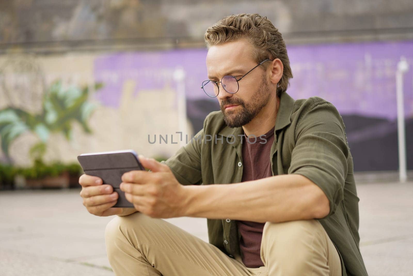 Mid-aged man engaged in studying from a tablet PC while sitting outdoors. With a focused expression, he utilizes the digital device to access study materials and engage in self-improvement. High quality photo