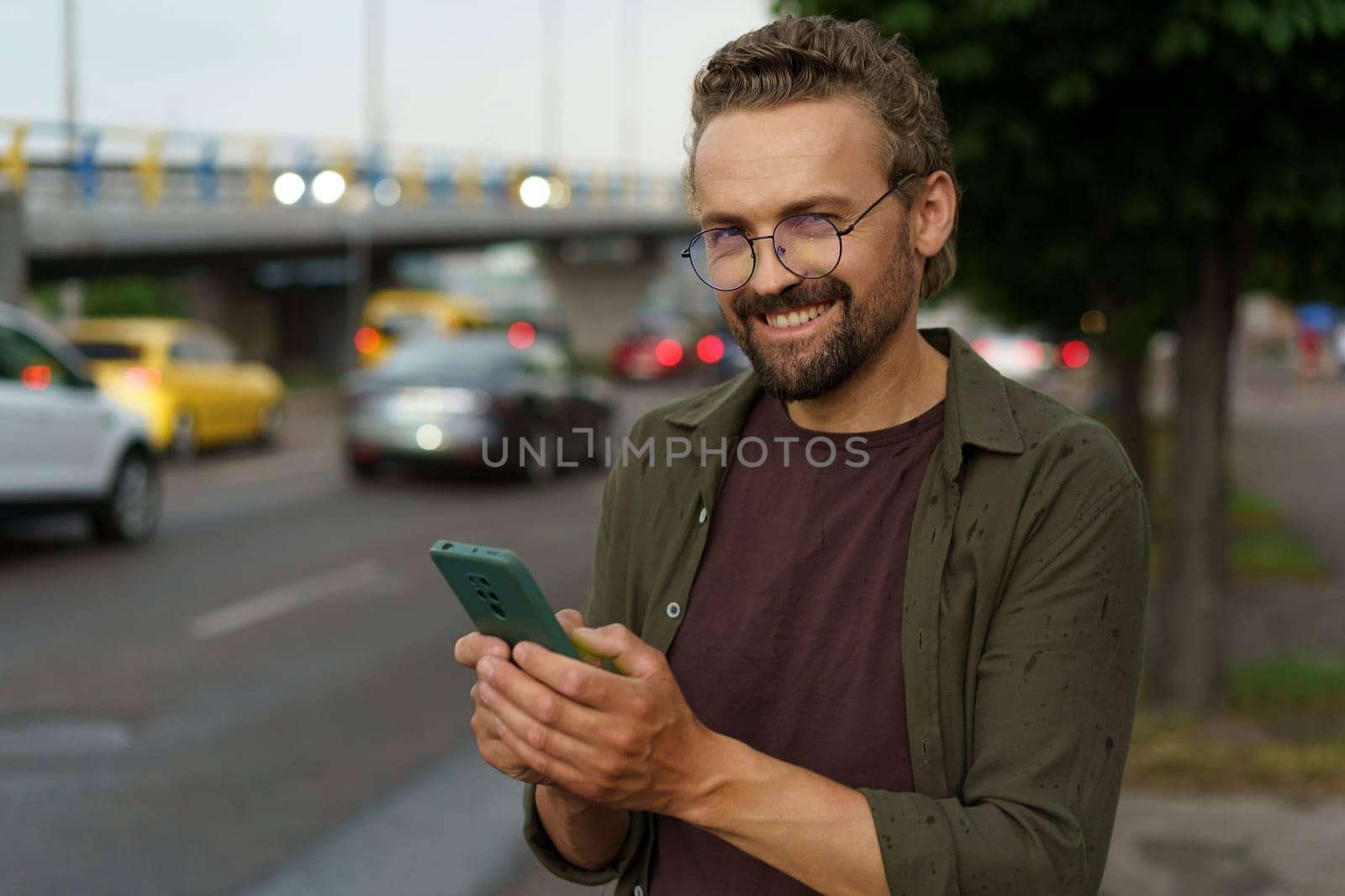 Happy customer making order for taxi cab. With smile on face, man uses phone to book ride while dusk sets in and street lights illuminate city. Presence of cars signifies bustling nature of city.