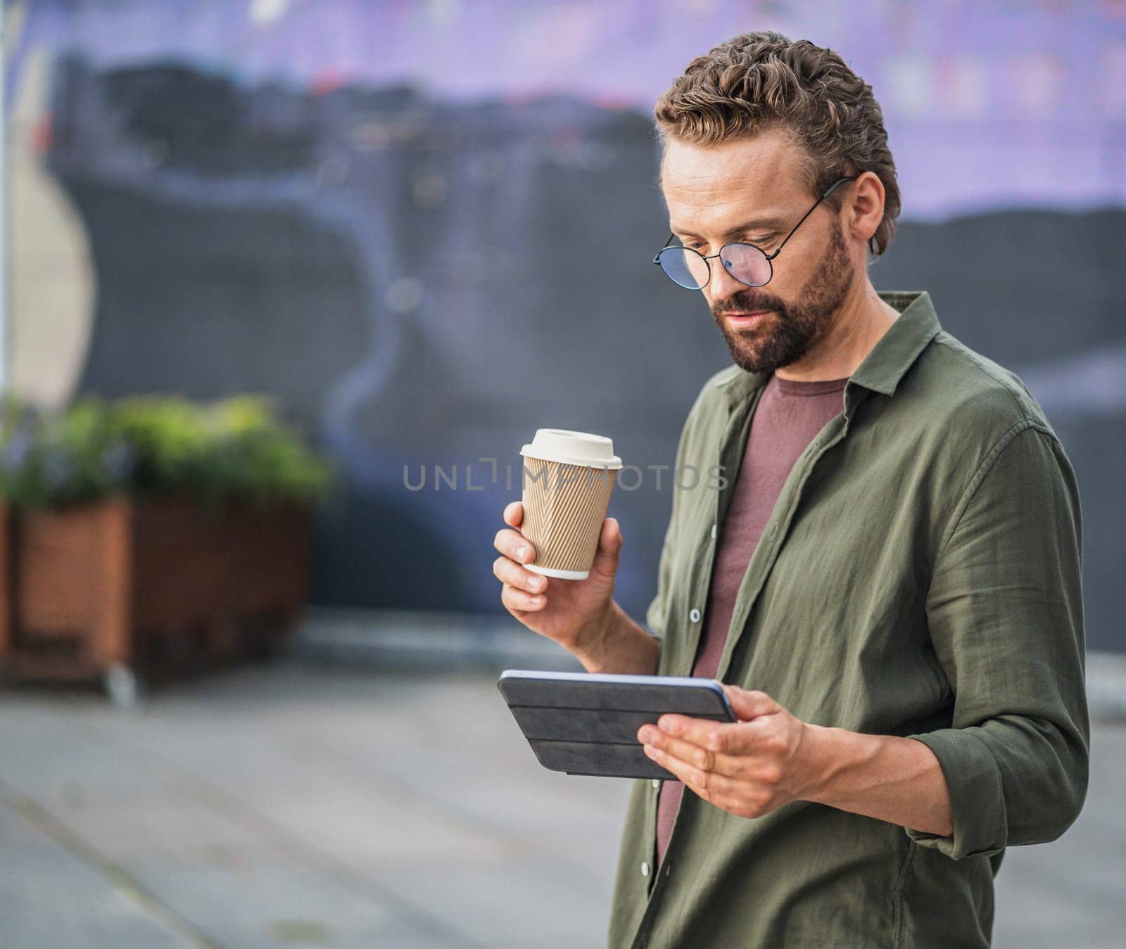 Man enjoying a coffee break outdoors while reading information from his tablet PC. The relaxed and serene atmosphere complements the combination of coffee and digital communication. by LipikStockMedia