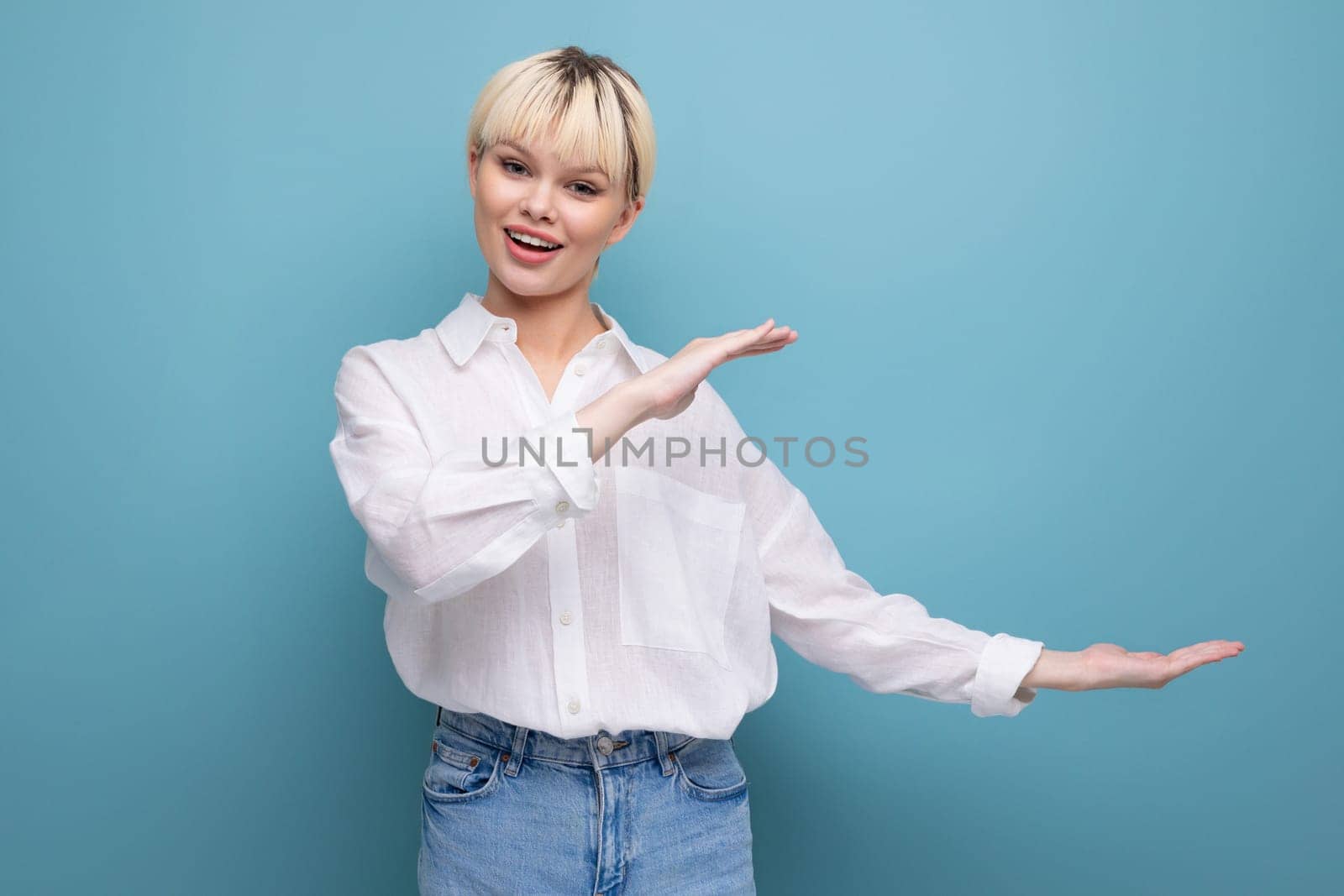 young blond caucasian woman with a short haircut dressed in a white shirt gesticulates with her hands and points to an empty space by TRMK