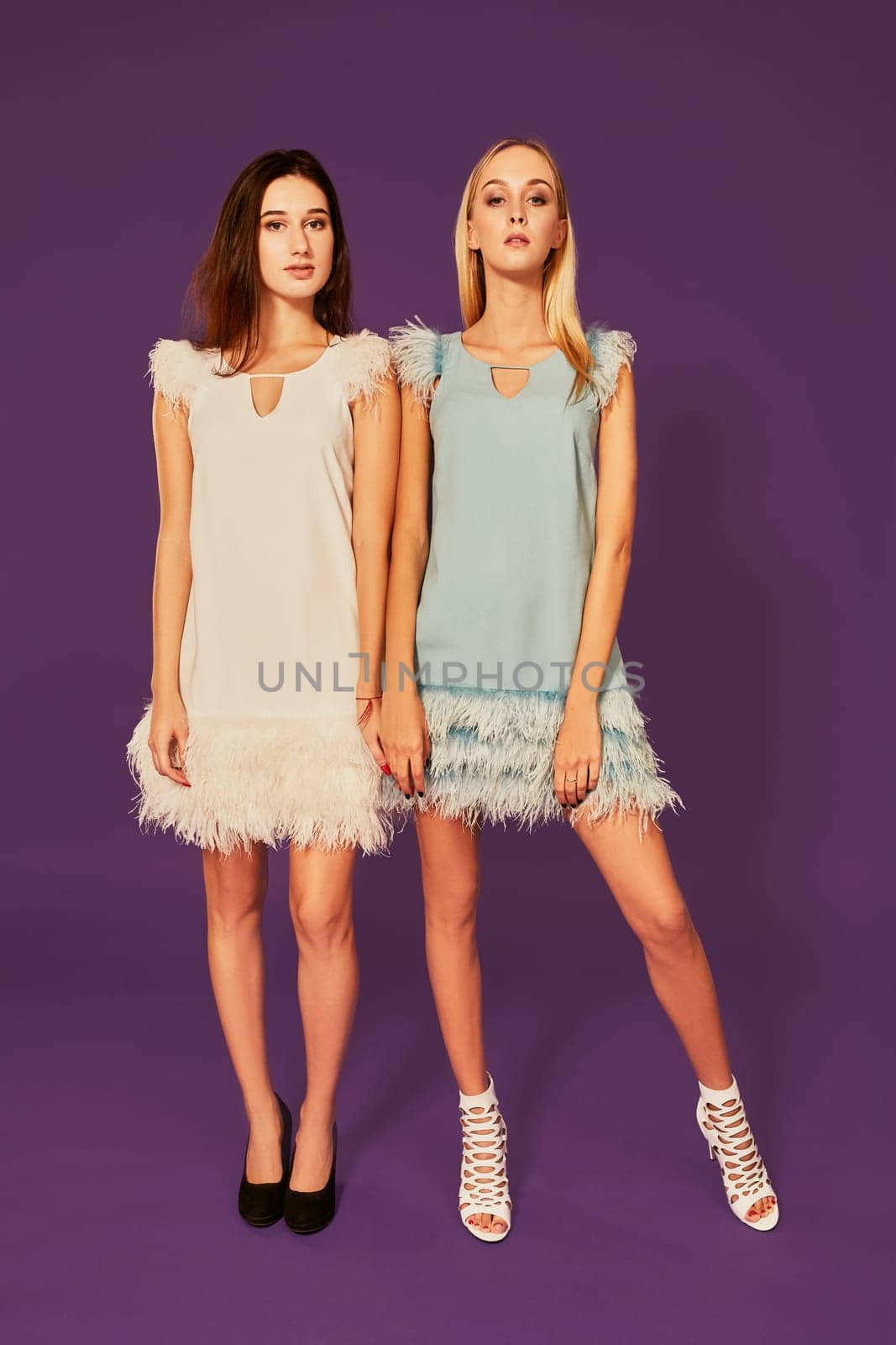 Professional photo of two young women, blonde and brunette, in cocktail dresses posing on purlpe background by nazarovsergey