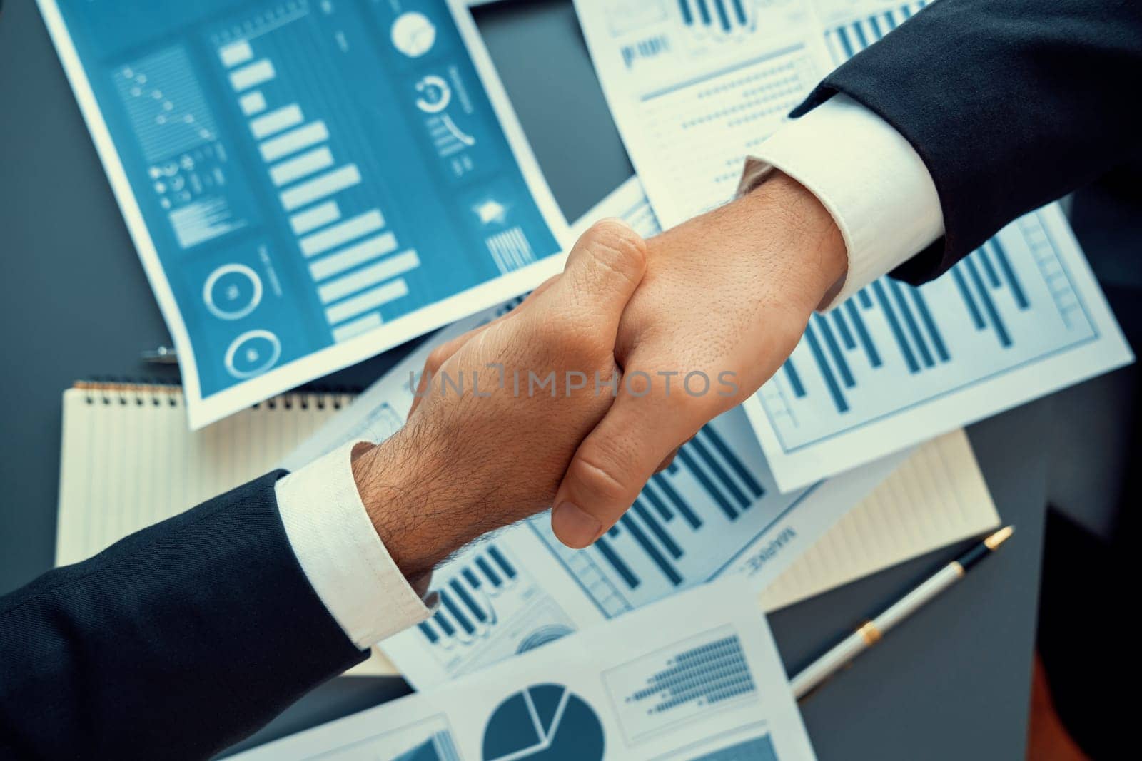Top view professional businessman shaking hands over desk in modern office after successfully analyzing pile of dashboard data paper as teamwork and integrity handshake in workplace concept. fervent
