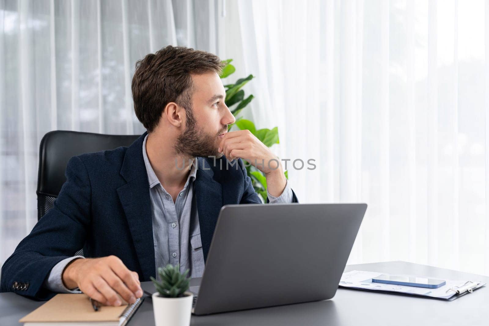 Businessman in black suit working on laptop at his workspace desk. Smart executive researching financial data and planning marketing strategy on corporate laptop at modern workplace. Entity