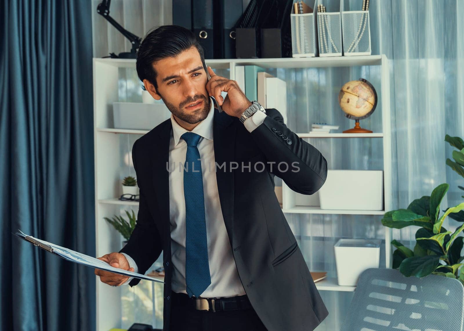 Diligent and busy businessman talking on phone call with coworker in modern office. Making sales call to client with coffee in hand, standing posing confidently as professional office worker. Fervent