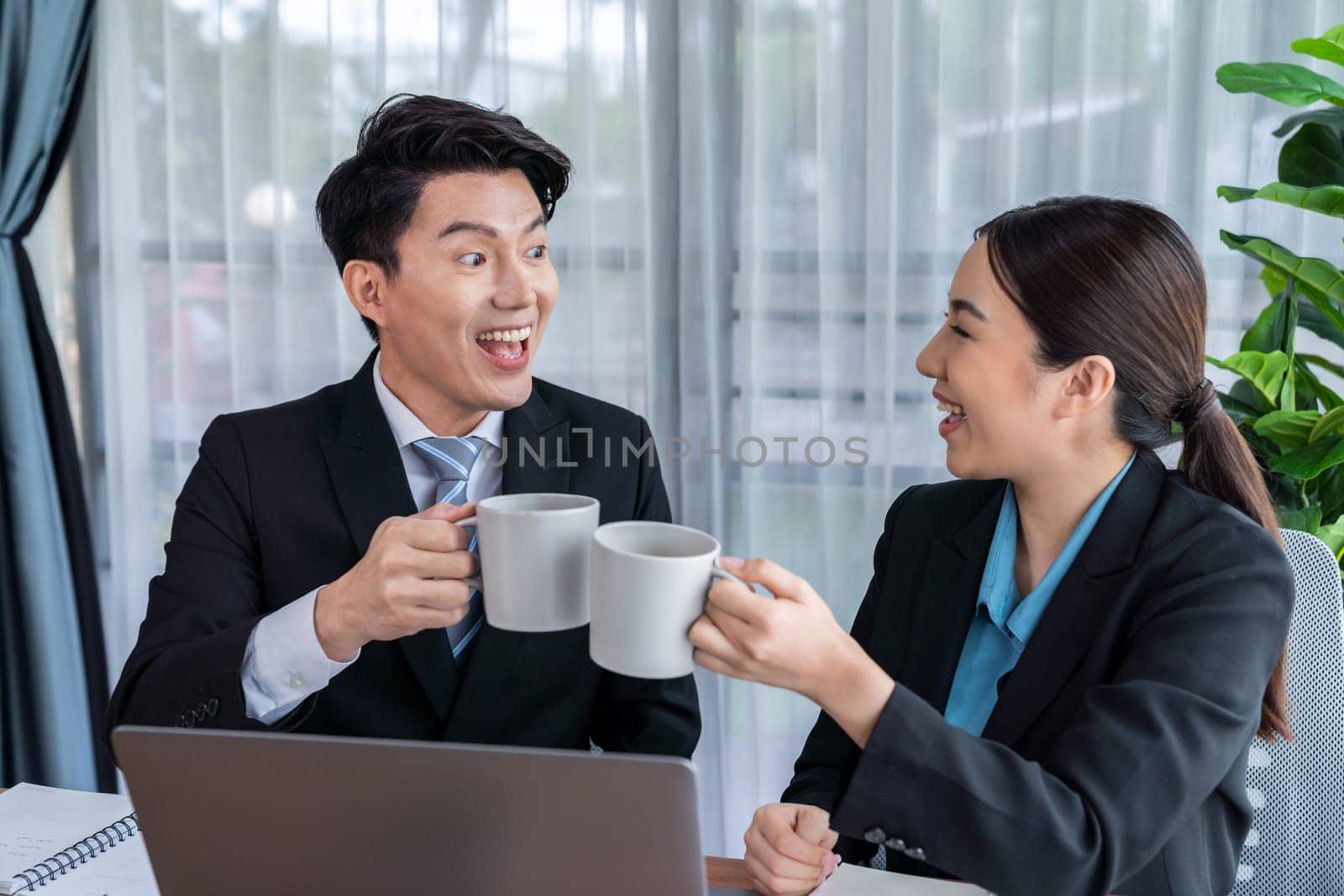 Two Asian office workers taking coffee break together in workplace. Coworkers smiling and socializing while holding cup of coffee adding friendly working environment in corporate workspace. Jubilant