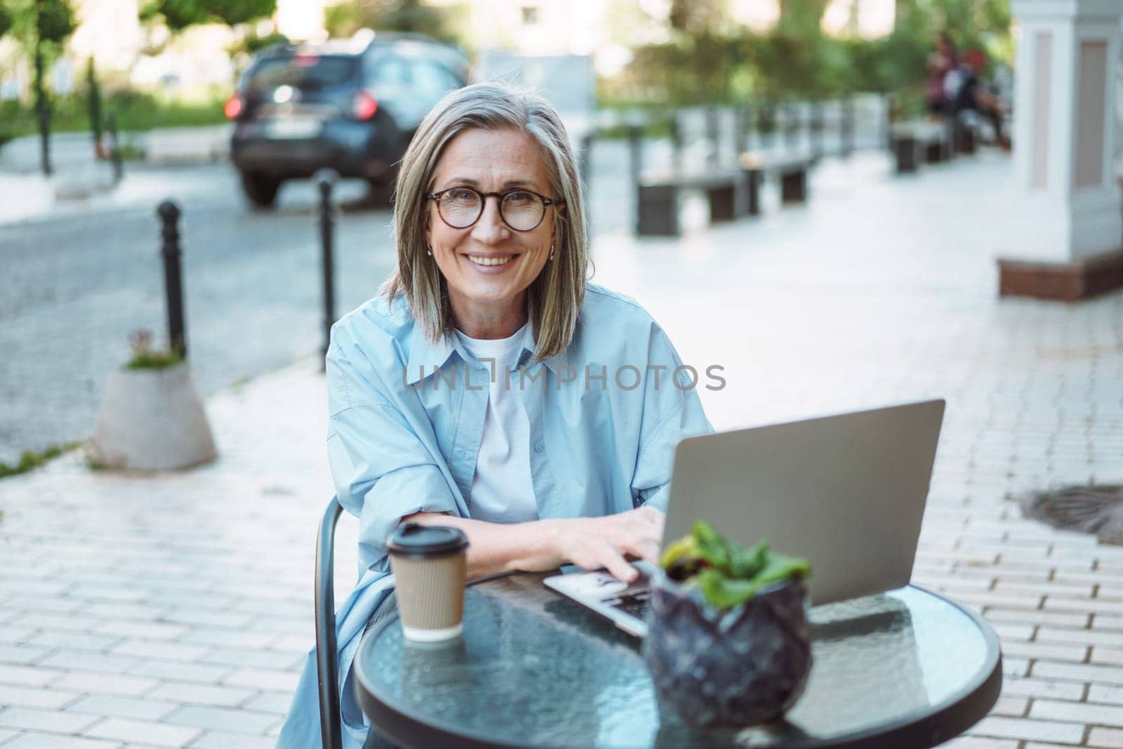 Working from anywhere, smiling and happy mature woman working on laptop from cafe on street. Freedom and flexibility that comes with remote work, allowing individuals to choose their work environment. by LipikStockMedia