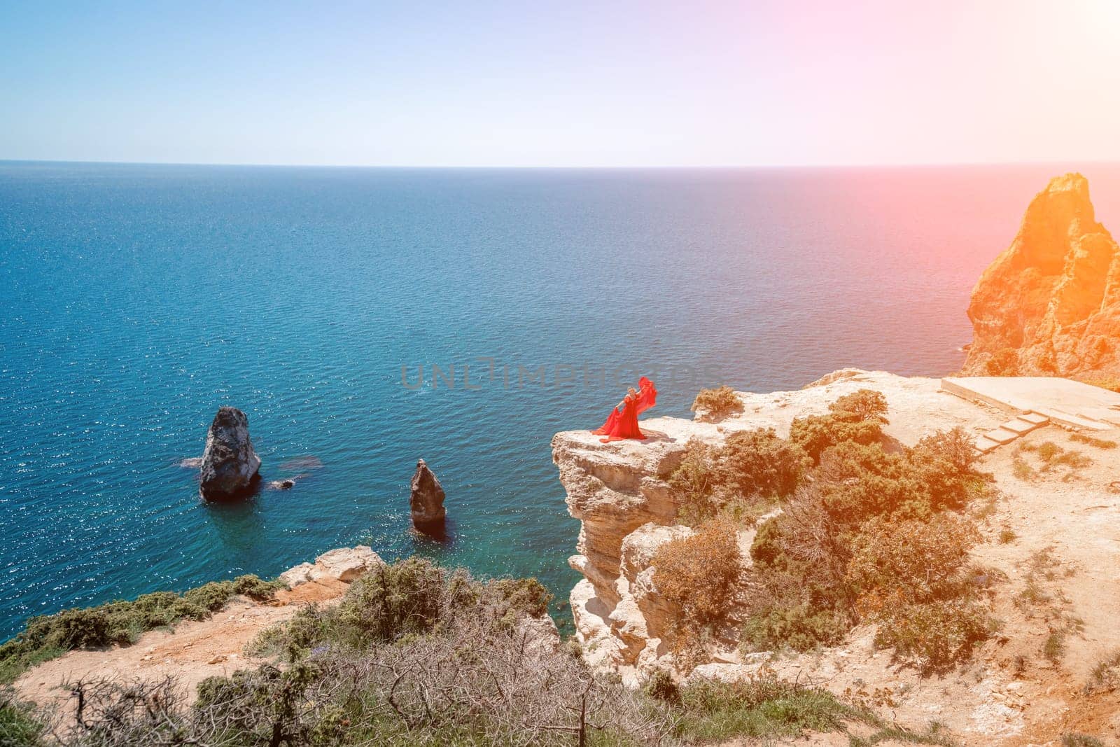 woman sea red dress. Blonde with long hair on a sunny seashore in a red flowing dress, back view, silk fabric waving in the wind. Against the backdrop of the blue sky and mountains on the seashore