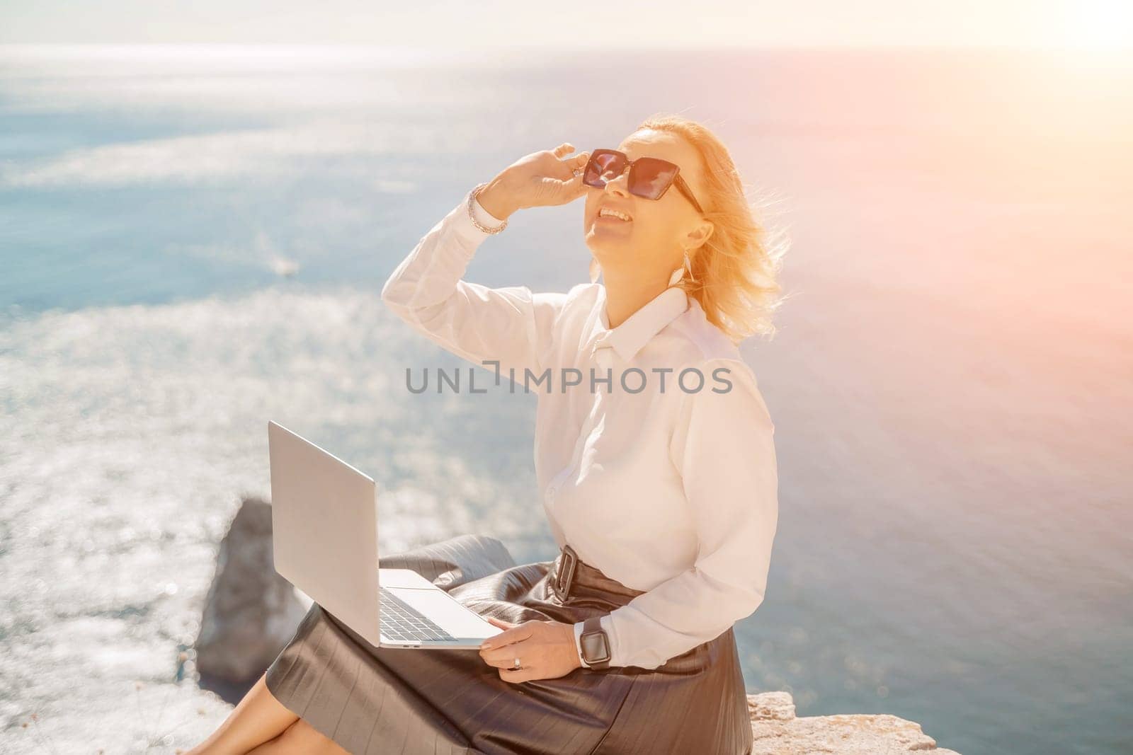 Business woman on nature in white shirt and black skirt. She works with an iPad in the open air with a beautiful view of the sea. The concept of remote work. by Matiunina