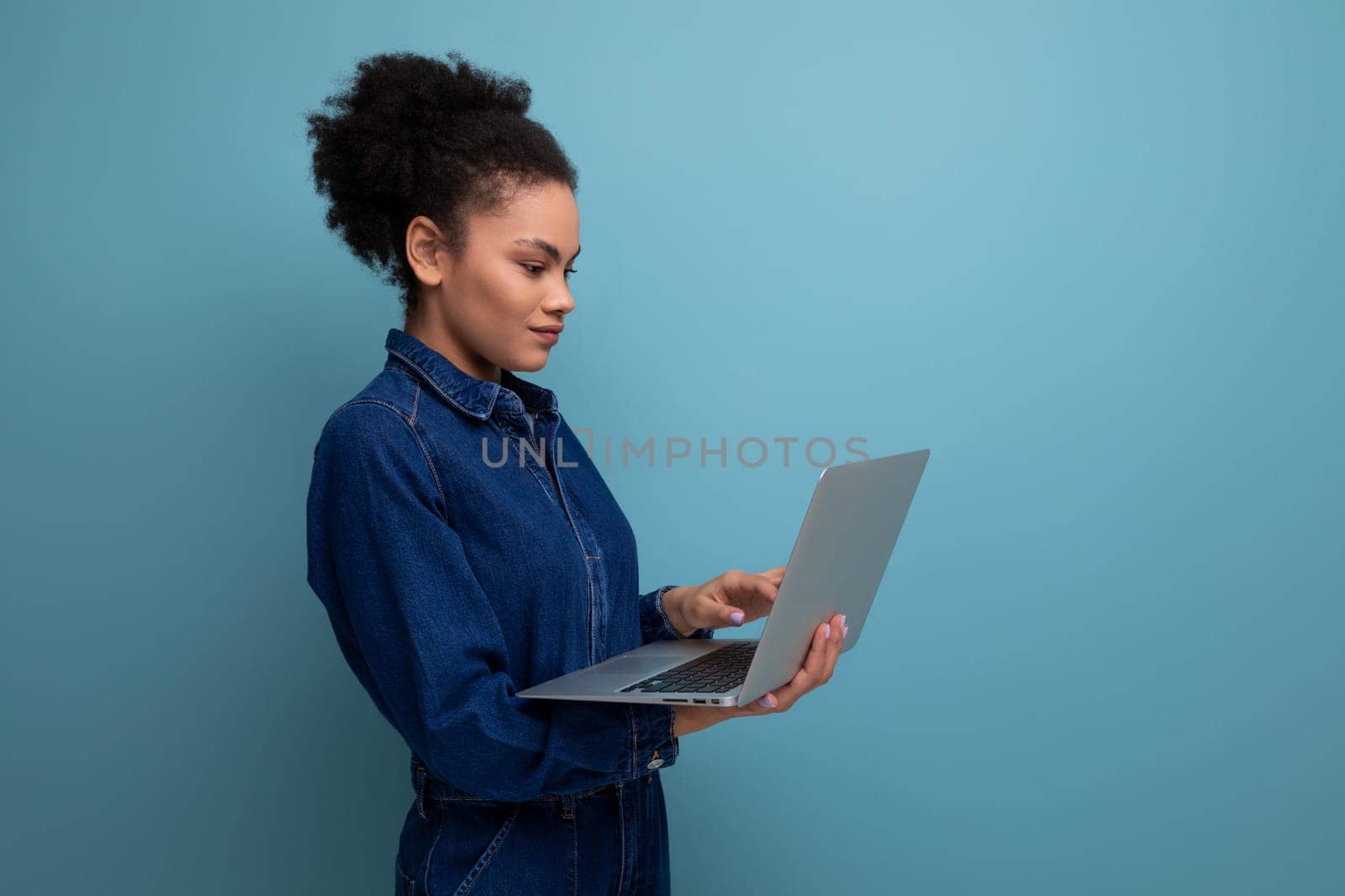young successful woman with dark skin and black curly hair dressed in a blue denim suit works remotely using a laptop.