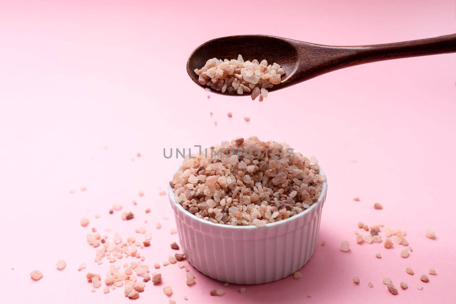 Pink Himalayan Rock Salt, Halite Pours From Wooden Spoon Over White Ceramic Bowl On Pink Background. Top View Horizontal Plane, Copy Space For Text.