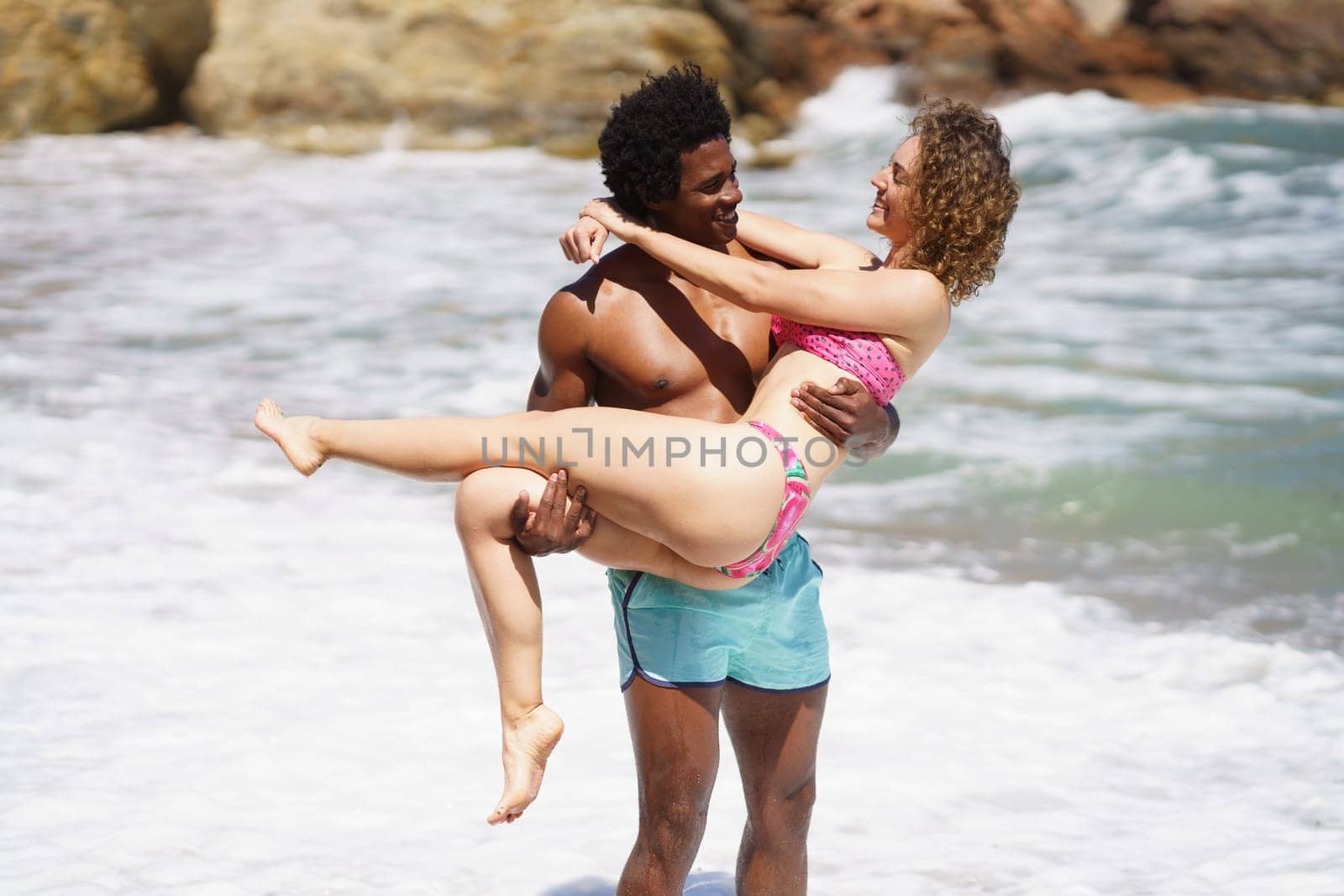 Smiling young multiracial couple in swimwear standing near waving blurred sea water and looking at each other while African American male carrying girlfriend in hands