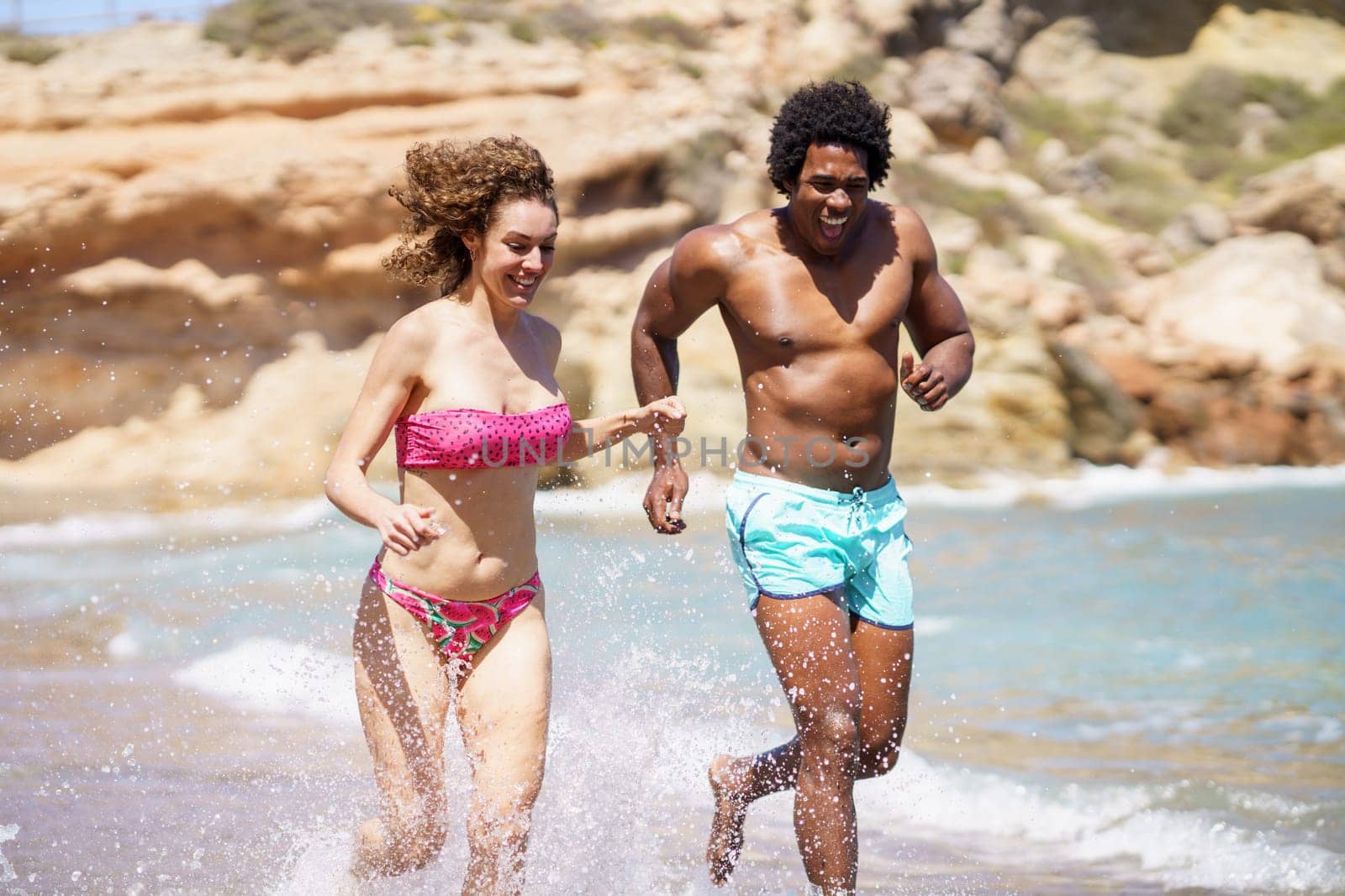 Young multiracial couple in swimwear running in splashing water while looking down and laughing together during sunny day near sea waves