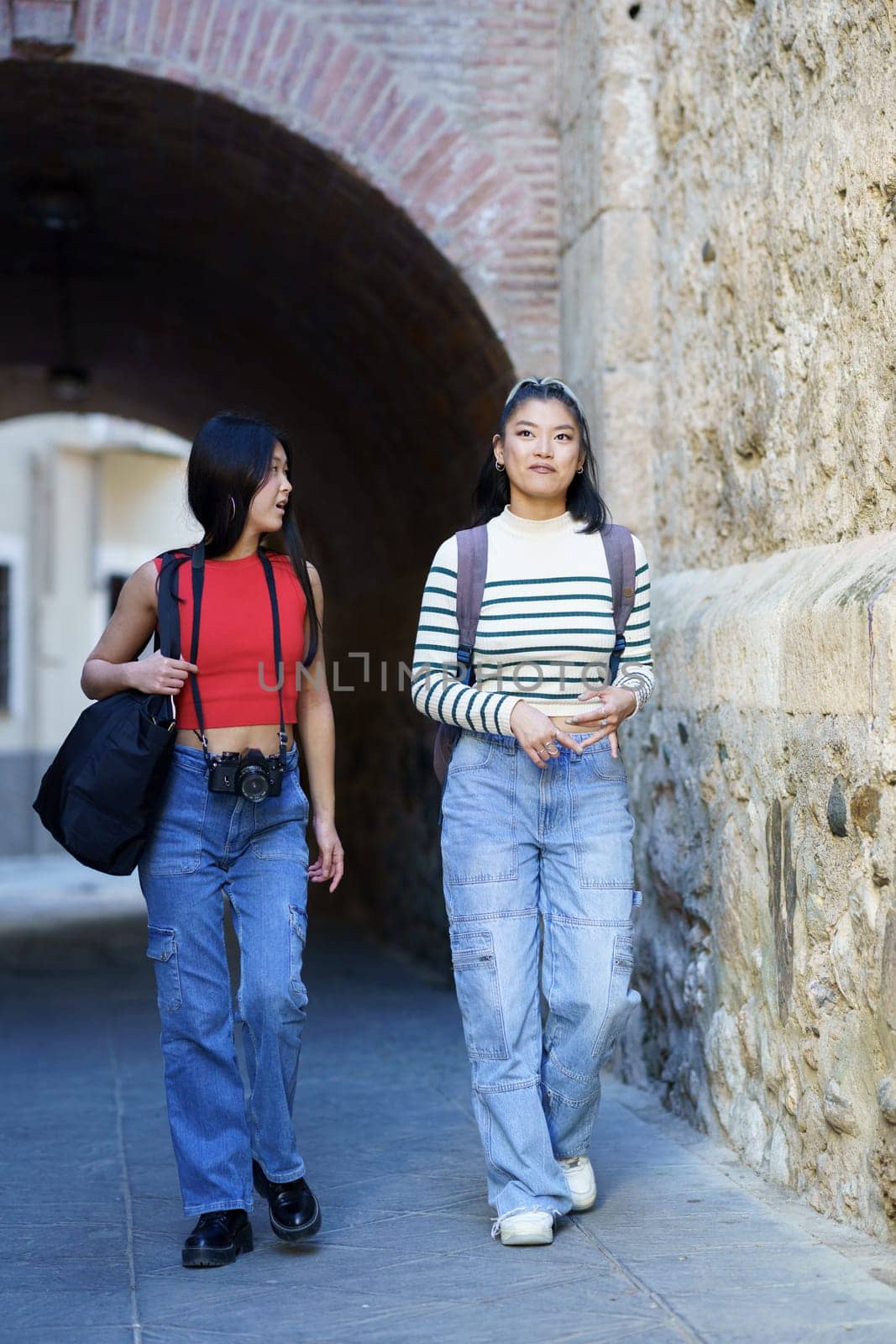 Full body of young Asian women tourists in casual clothes with bags and photo camera strolling against stone wall while talking to each other