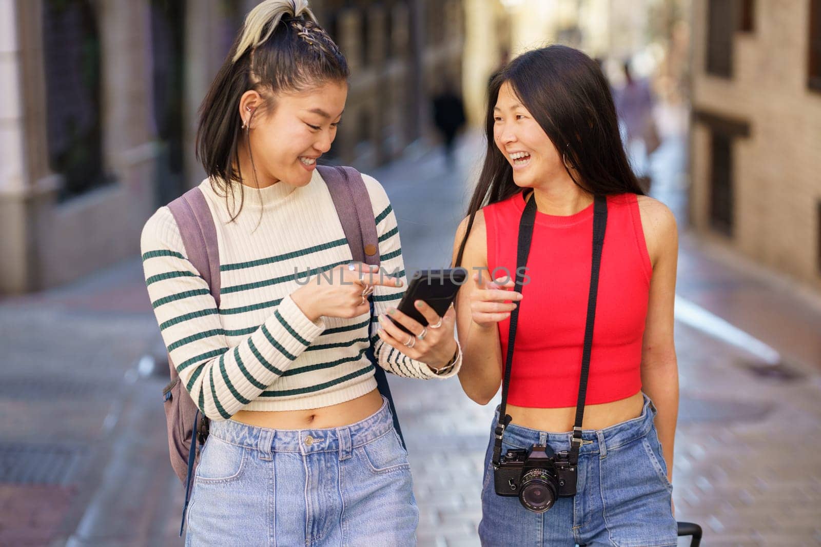 Positive young Asian women in casual clothes with backpack and photo camera, walking on town street while pointing at smartphone screen and laughing during trip through Spain